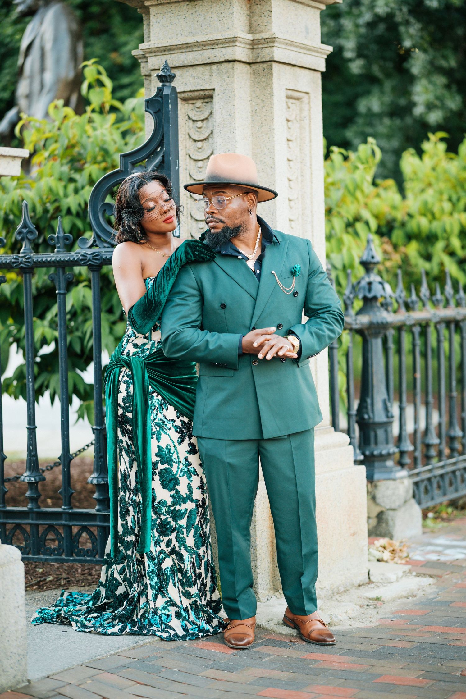 Elegant couple in coordinated green outfits posing by decorative iron gate and stone pillar.