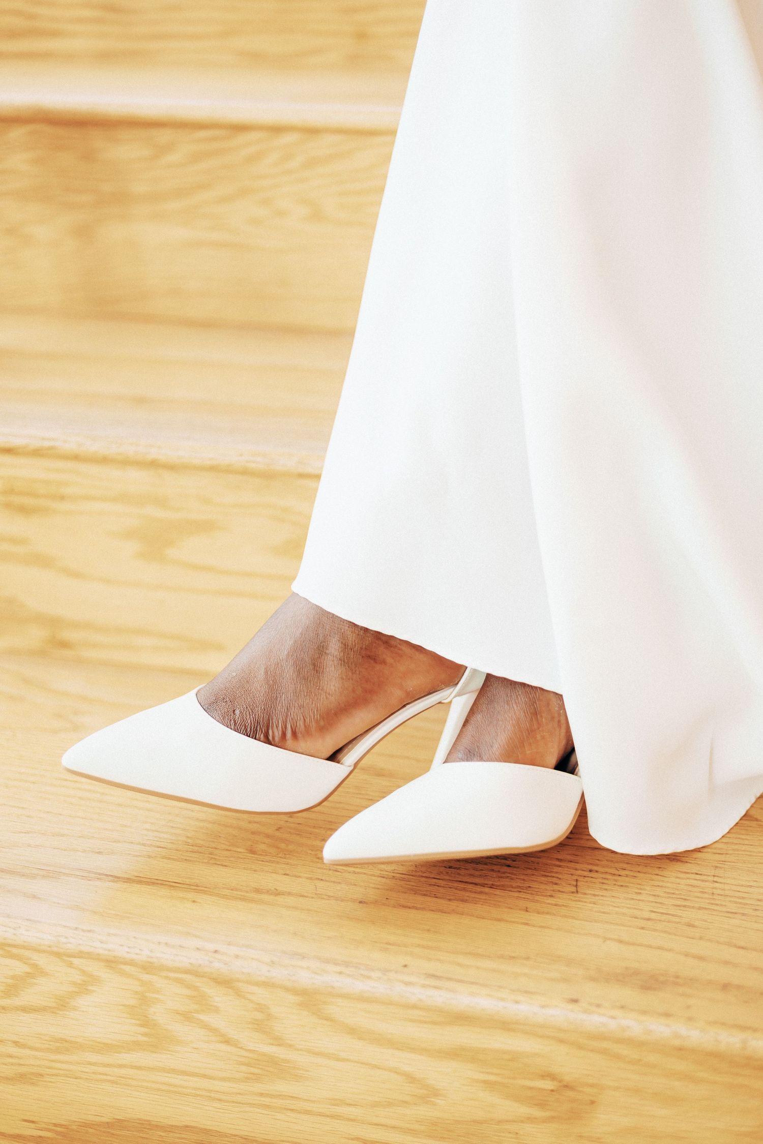 Close-up of a bride wearing white pointed-toe slingback heels and a flowing white gown on wooden stairs.