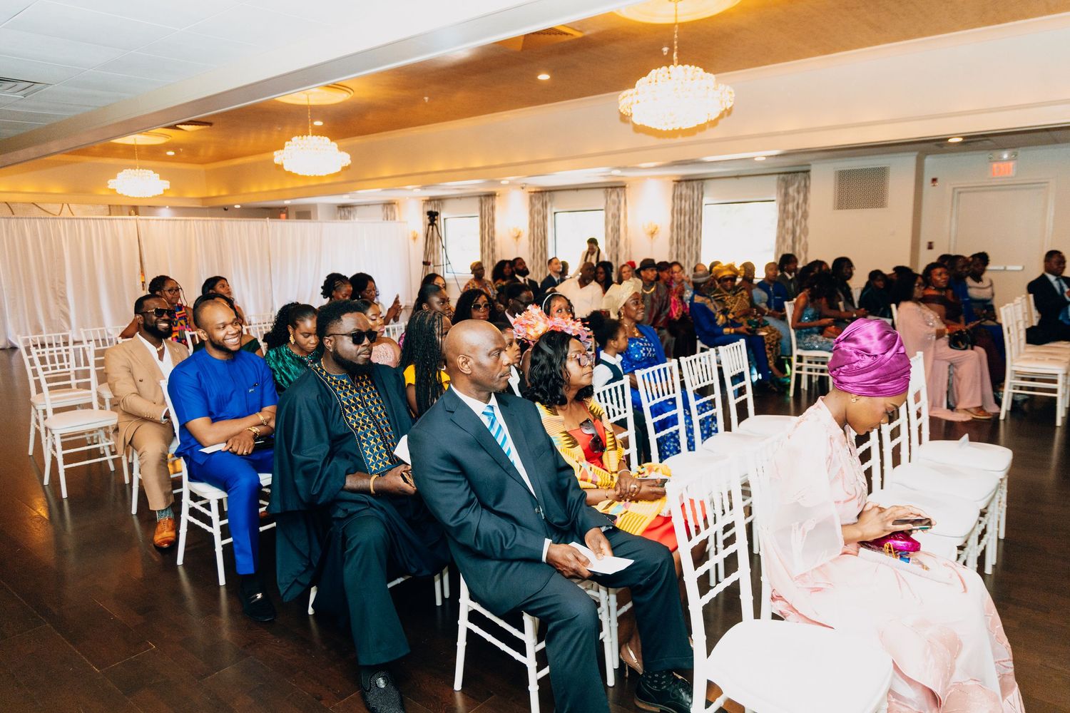 Guests seated in elegant white chairs at a formal indoor event in a decorated banquet hall.