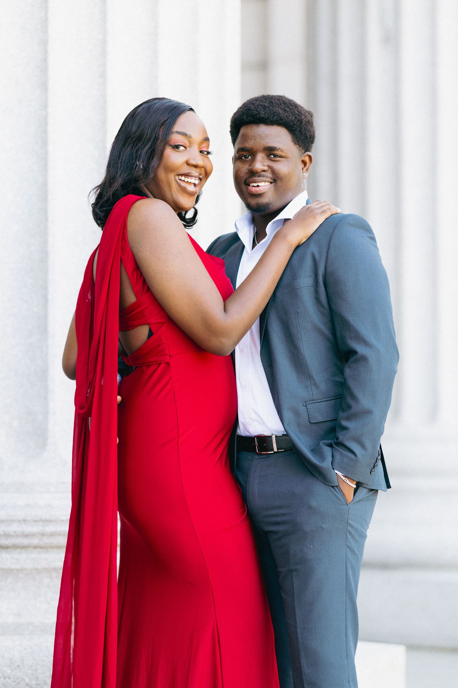 Smiling couple in formal attire, woman in red gown and man in gray suit, posing by stone pillars.