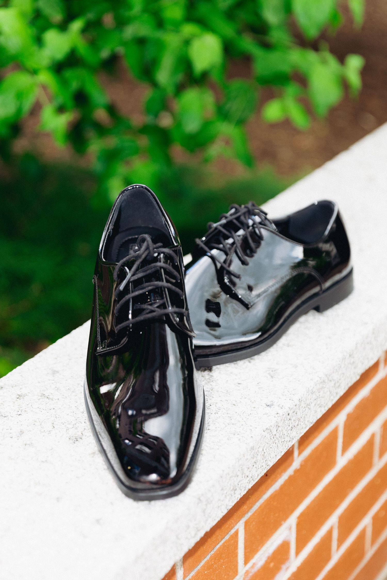 Shiny black patent leather Oxford dress shoes with laces displayed on a white stone ledge outdoors.