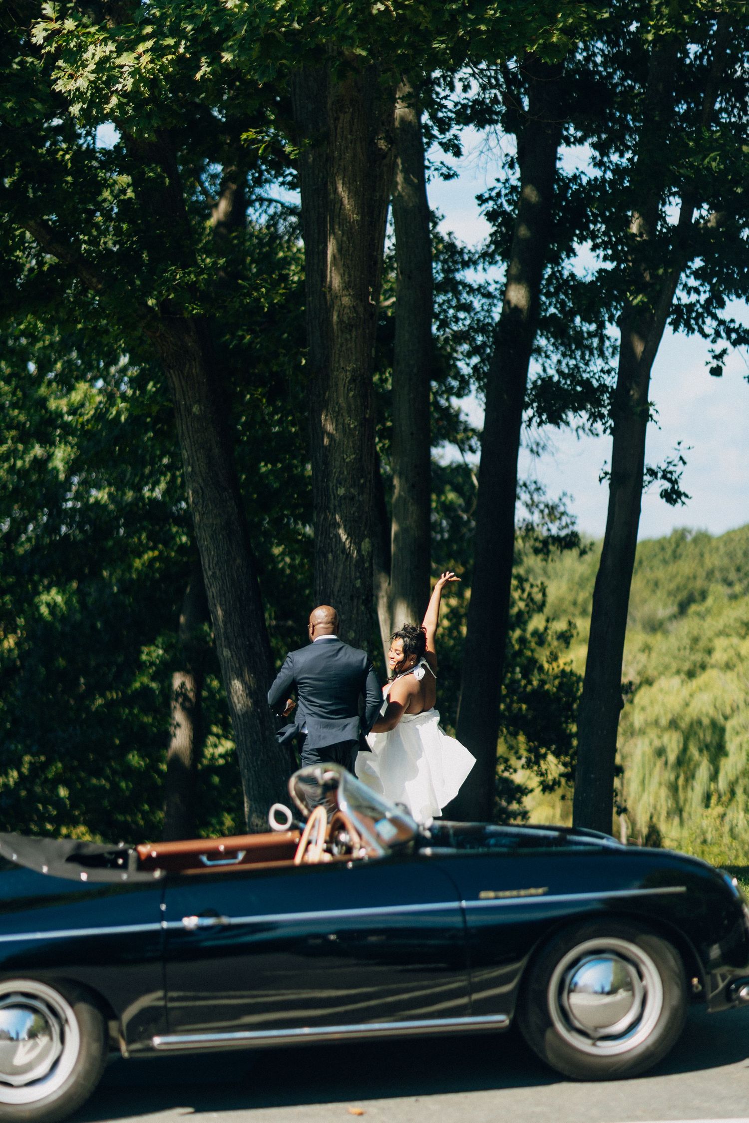 Bride and groom standing in classic black Porsche 356 convertible surrounded by tall green trees.