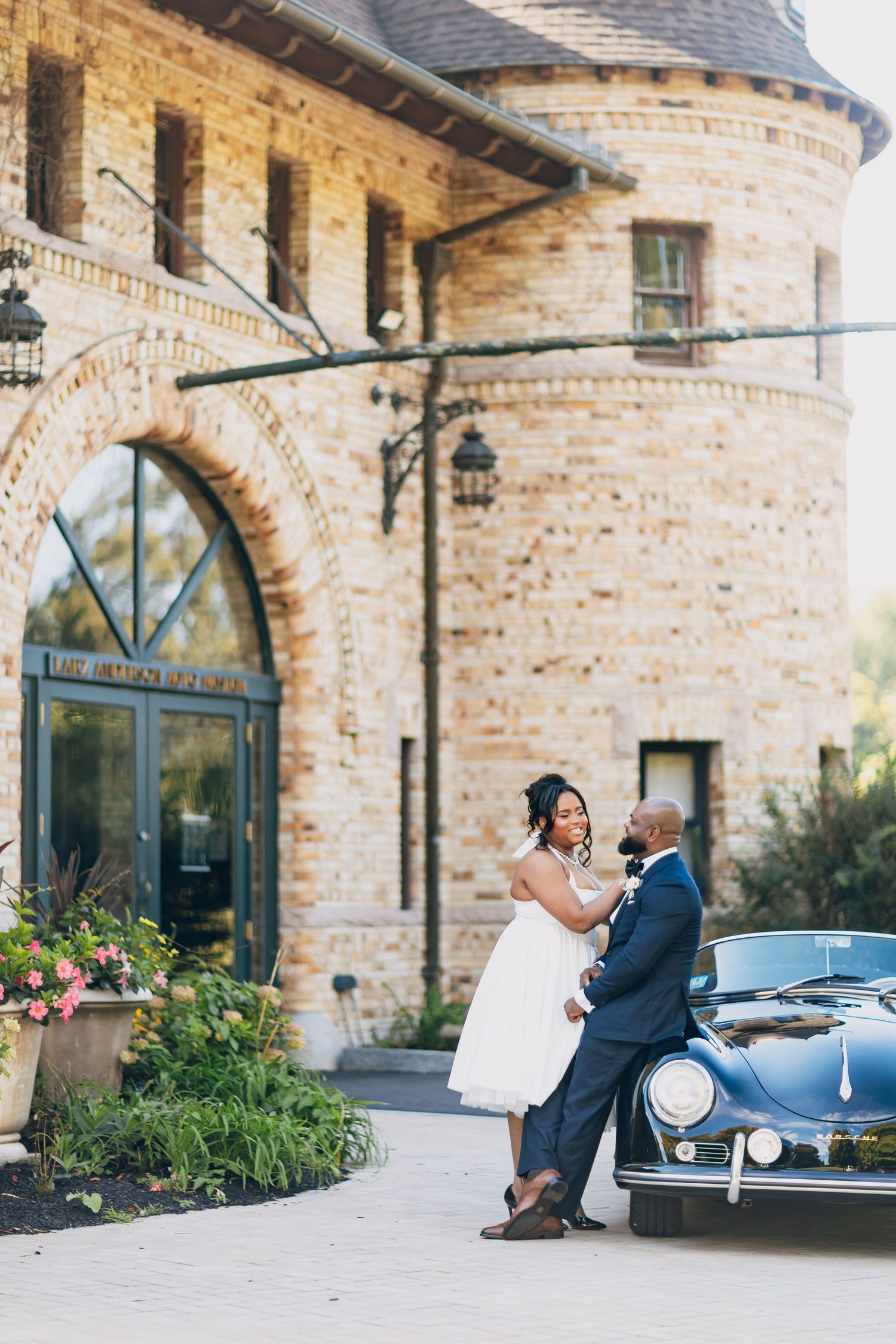 Bride and groom pose romantically beside a vintage black Porsche outside an elegant brick building.