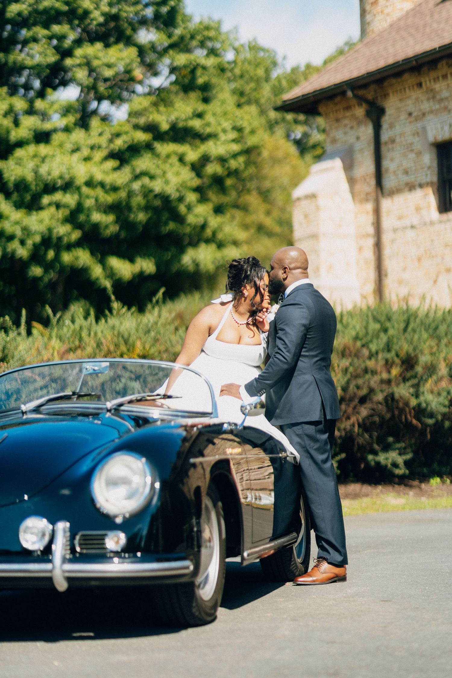 A bride and groom share a romantic moment leaning against a classic vintage black Porsche convertible outdoors.