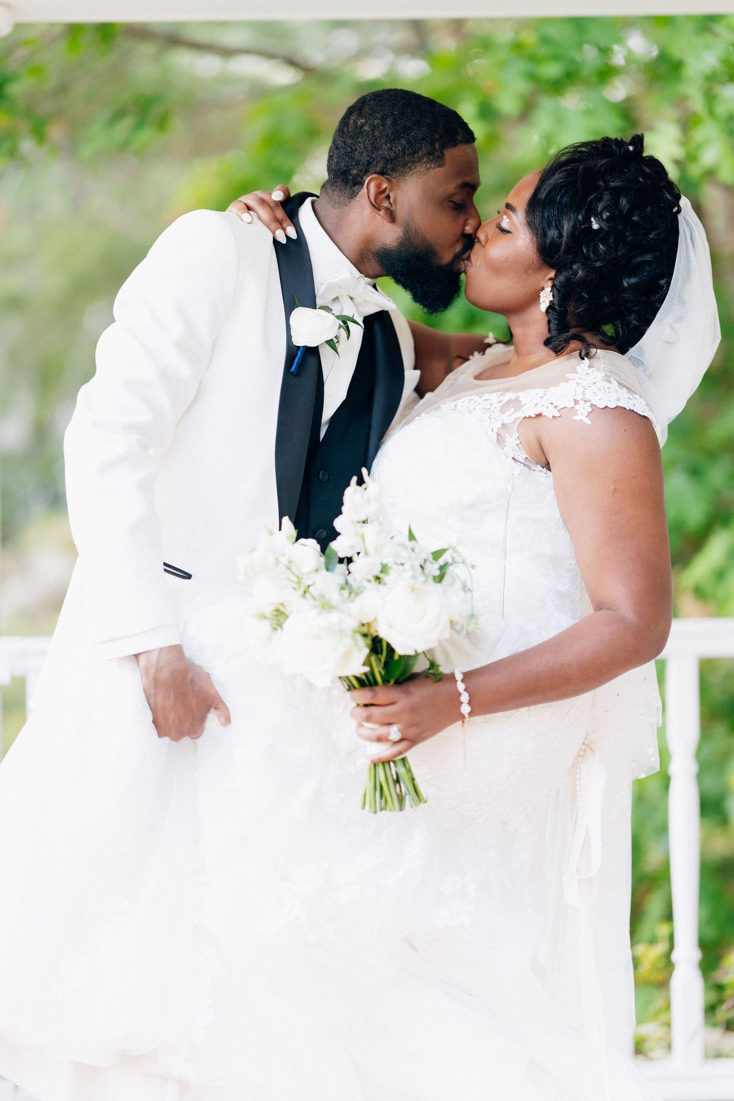 A Black bride and groom share a kiss at their wedding, bride holding white bouquet in lace gown with veil.
