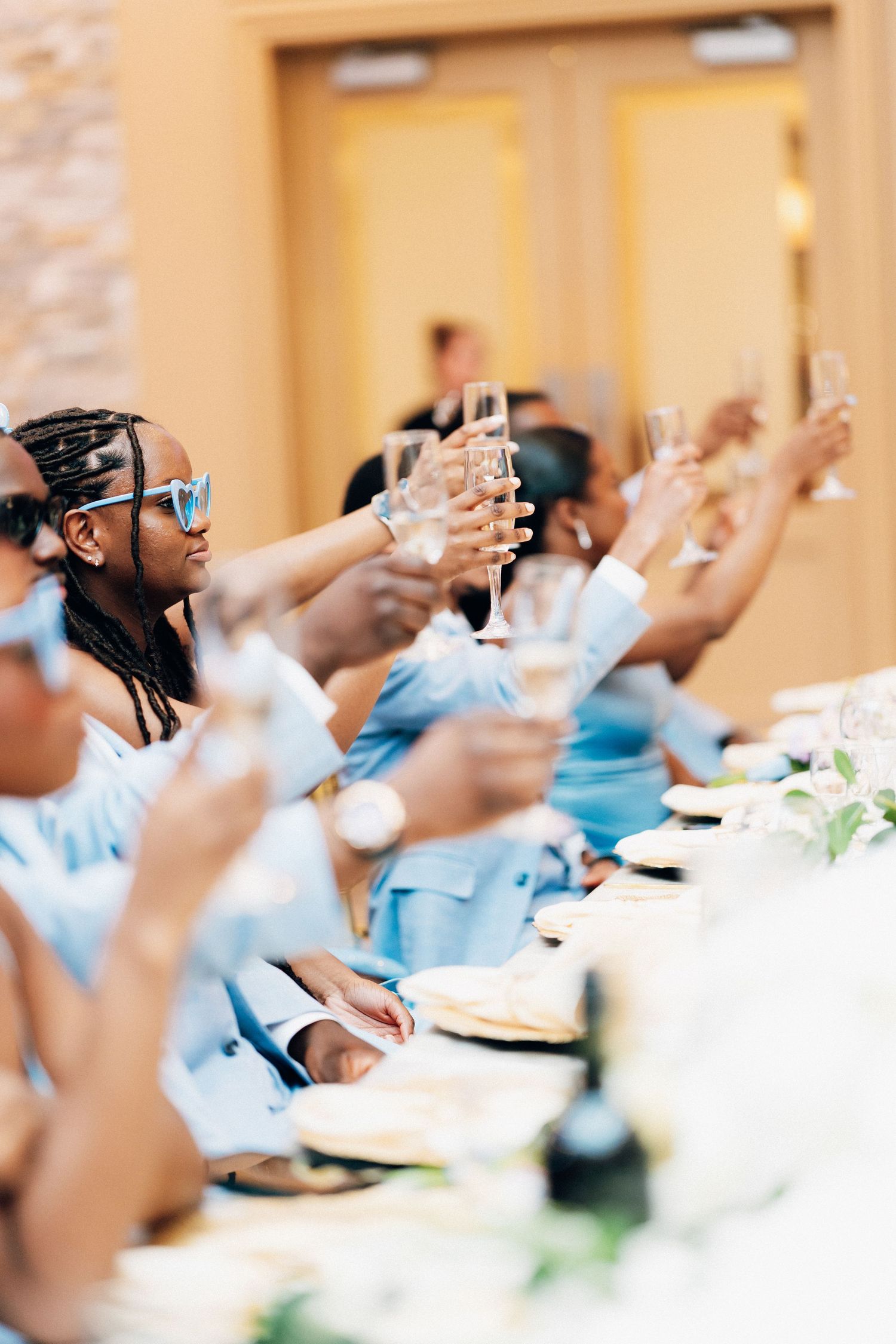 Wedding party in light blue attire raising champagne glasses in a toast at the reception table.