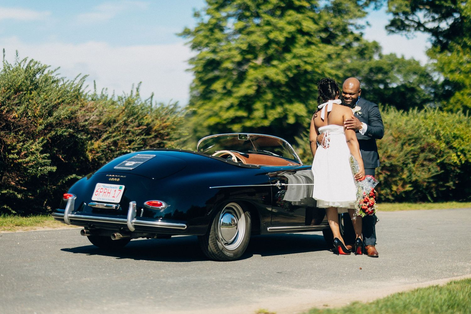 Black bride and groom embracing behind a classic navy blue vintage Porsche convertible on a sunny day.