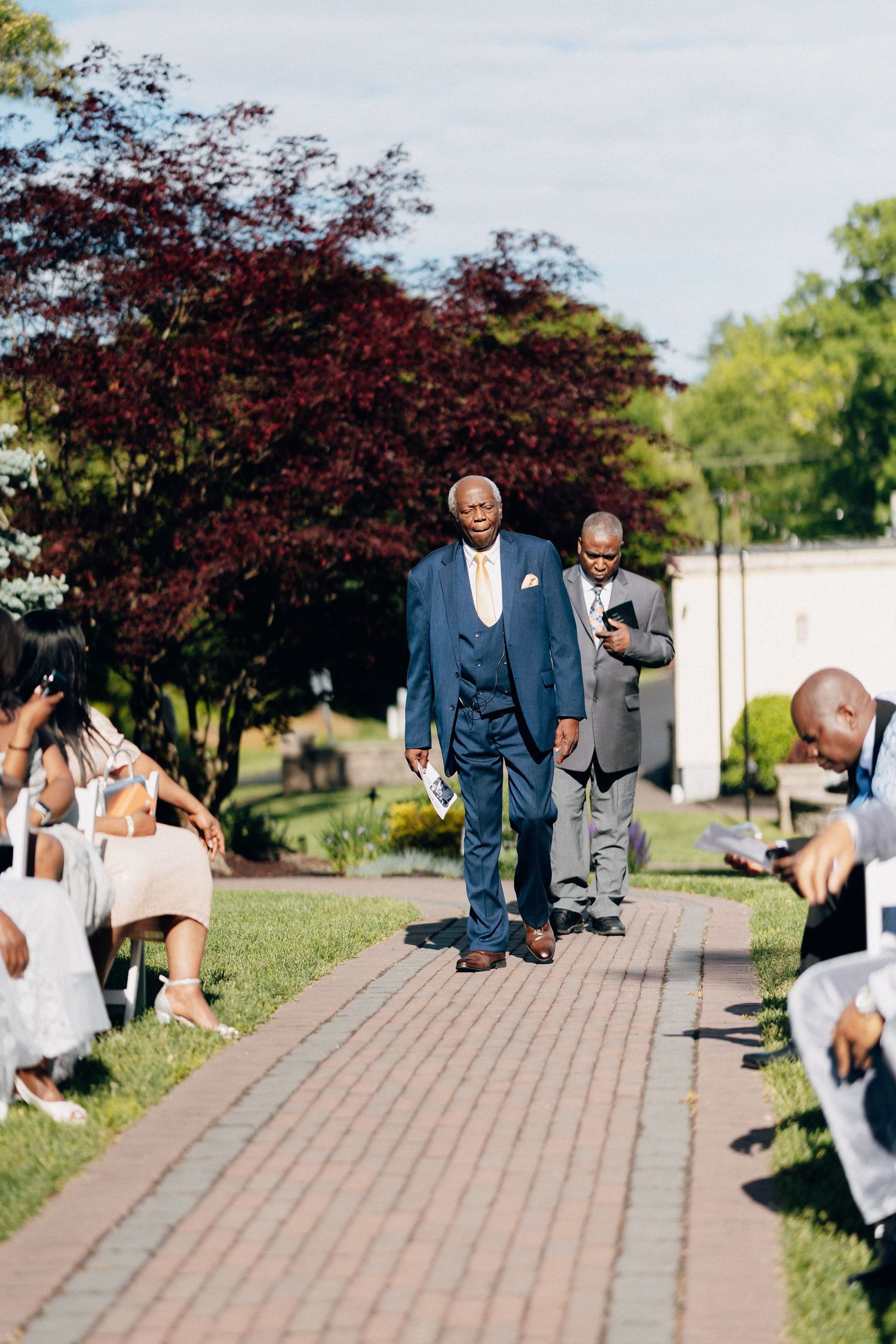 Elderly man in blue suit walks down outdoor wedding aisle lined with seated guests and lush green trees.
