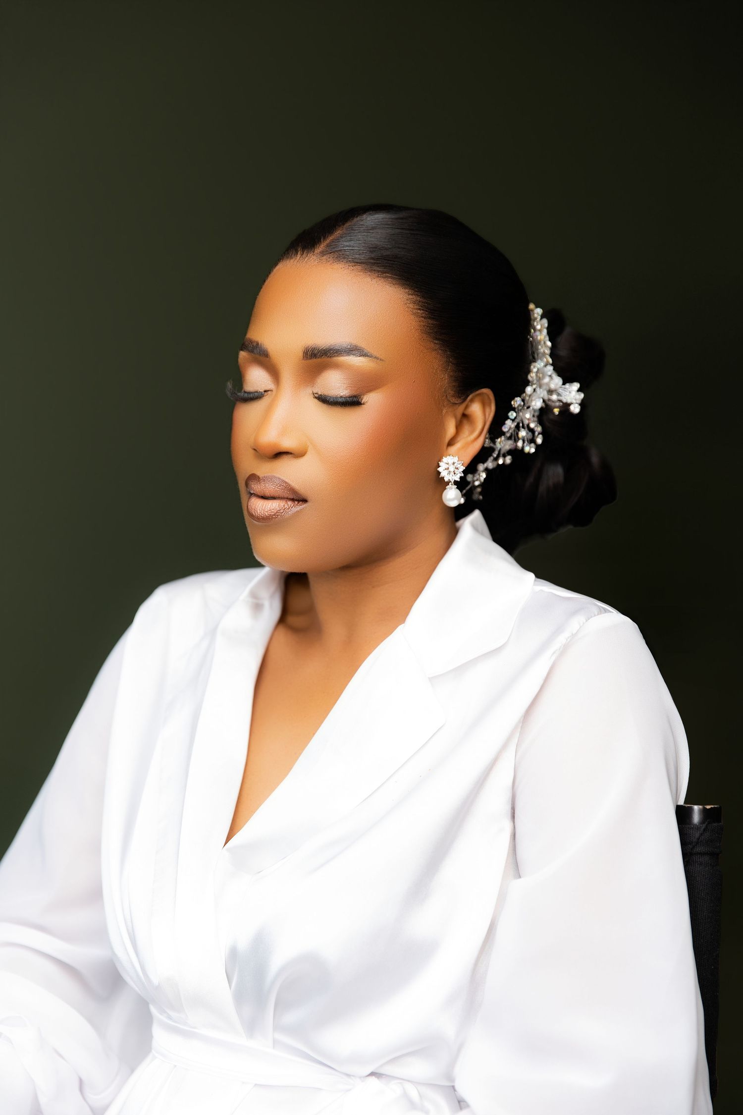 Elegant Black bride in white satin robe with pearl hair accessory, eyes closed, against dark green background.