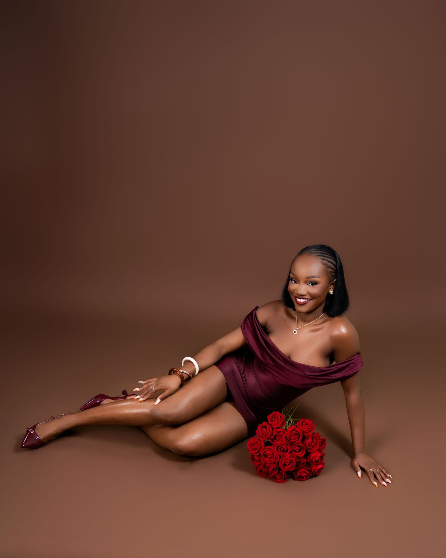 Woman in burgundy dress poses with red roses bouquet on brown studio backdrop.