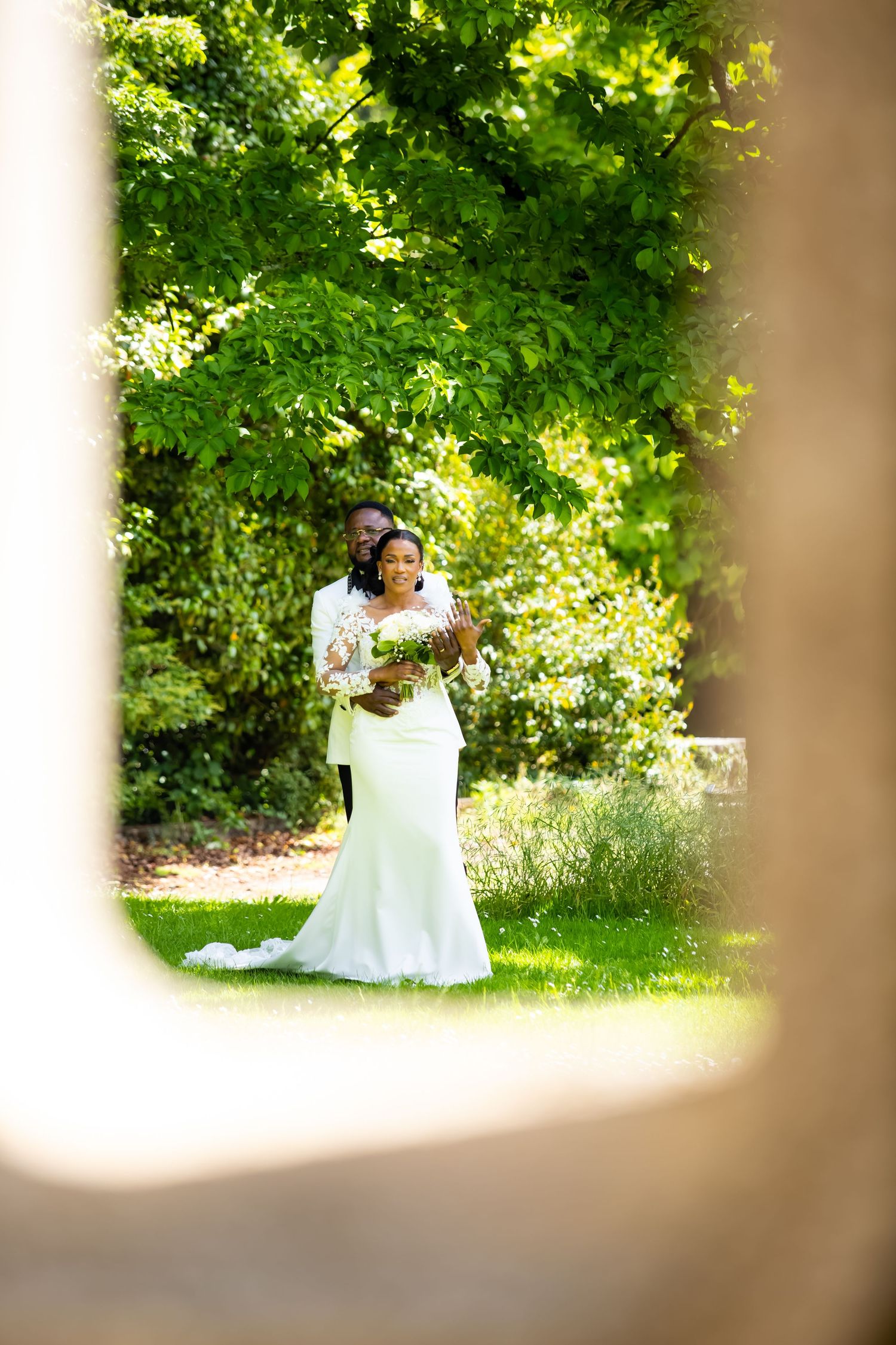 Black bride and groom in white wedding attire pose romantically in a lush green garden, framed by stone architecture.