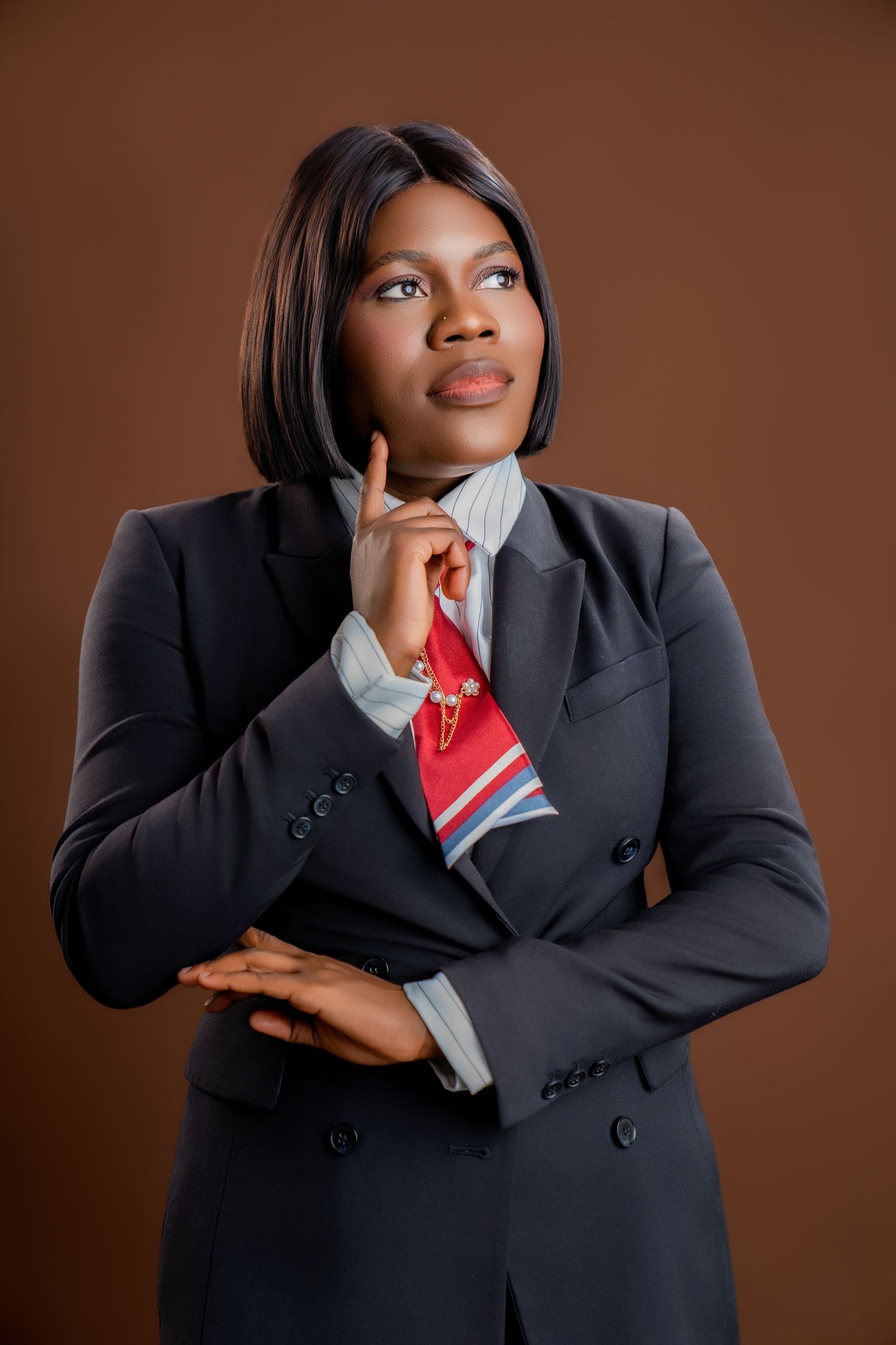 Professional Black woman in dark blazer and red tie, posing thoughtfully with finger raised against brown background.