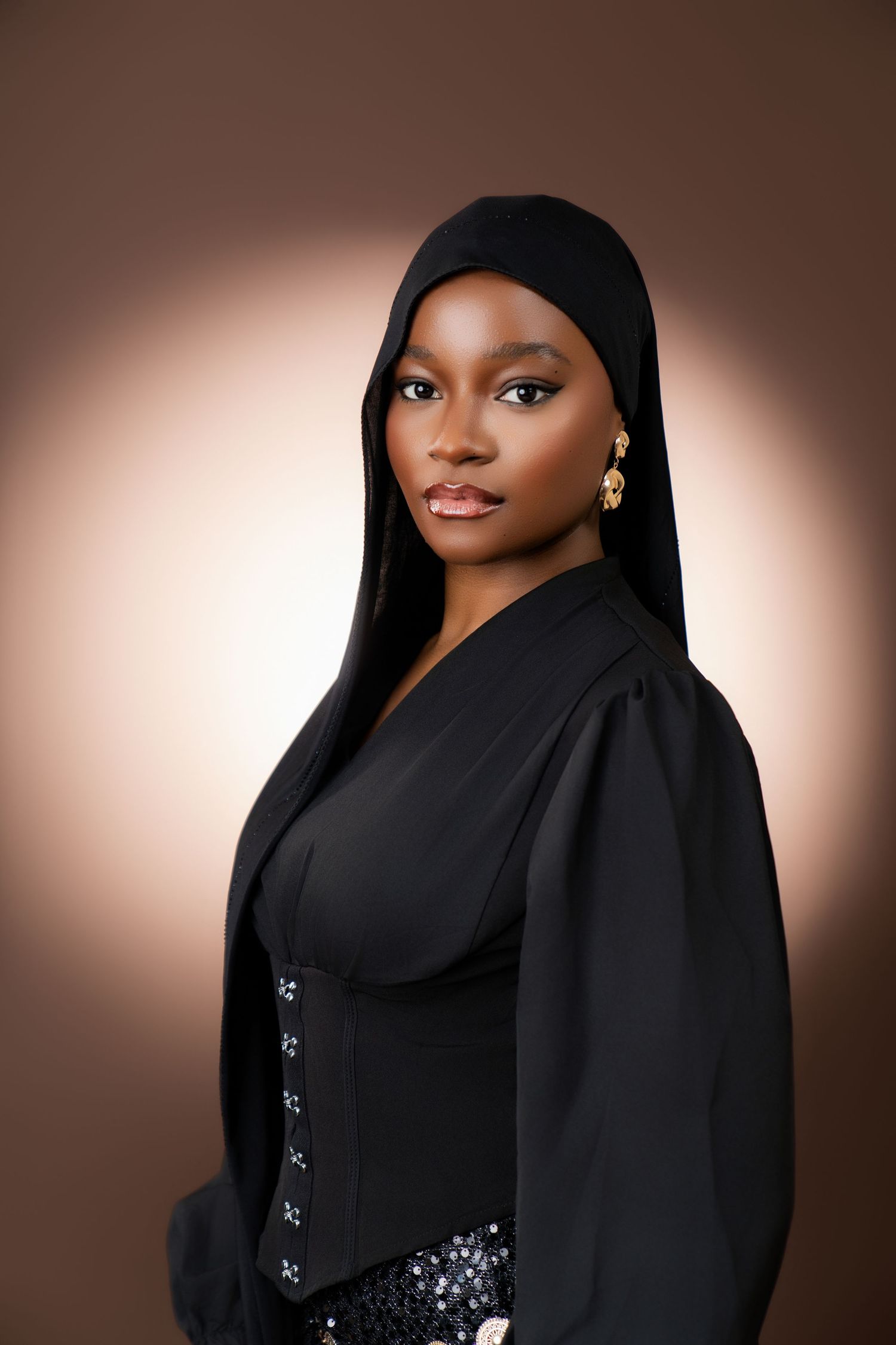 Young Black woman in black hijab and sequined outfit poses elegantly against warm brown studio background.