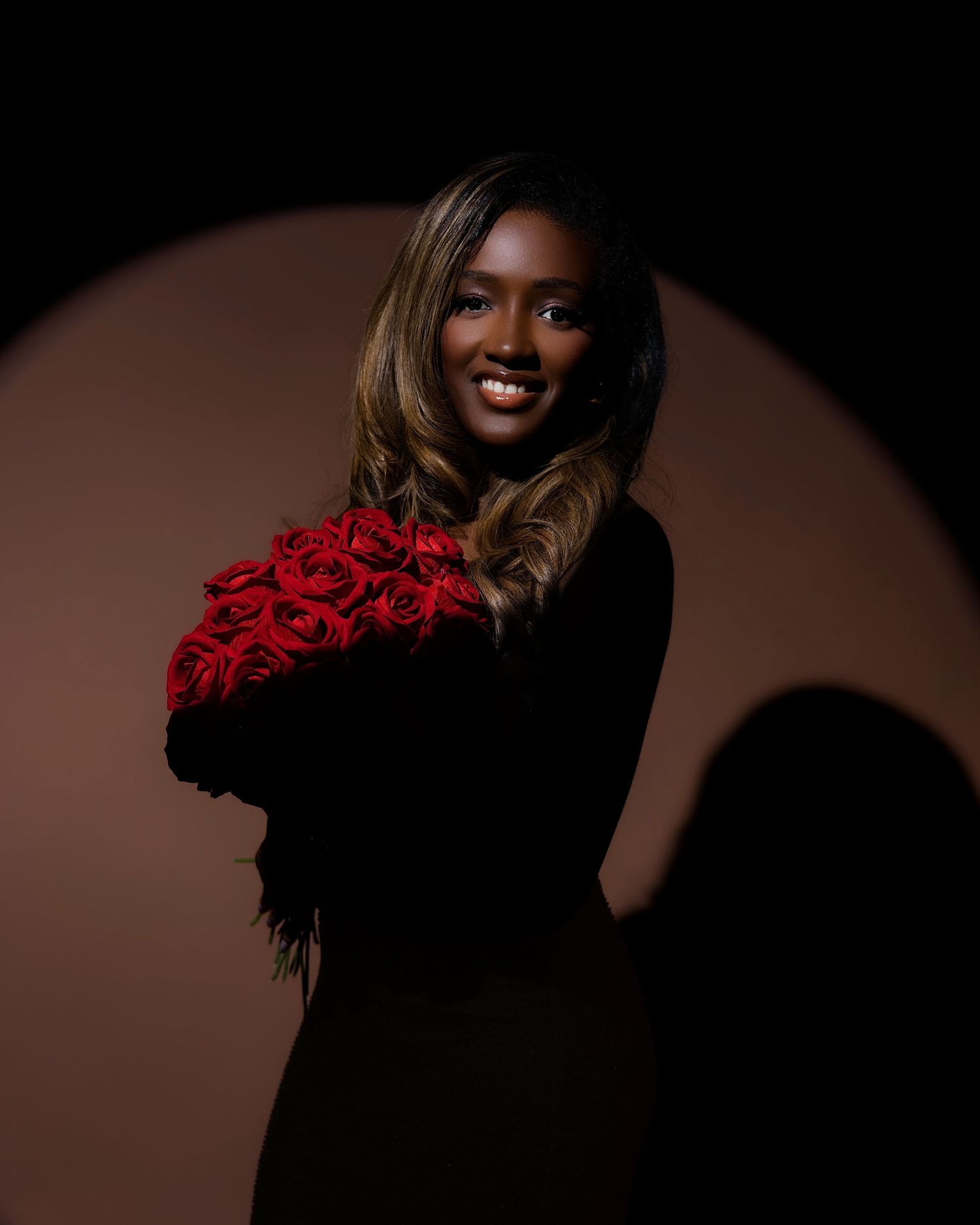 Woman in black dress smiling and holding red roses bouquet against dark background with circular light.