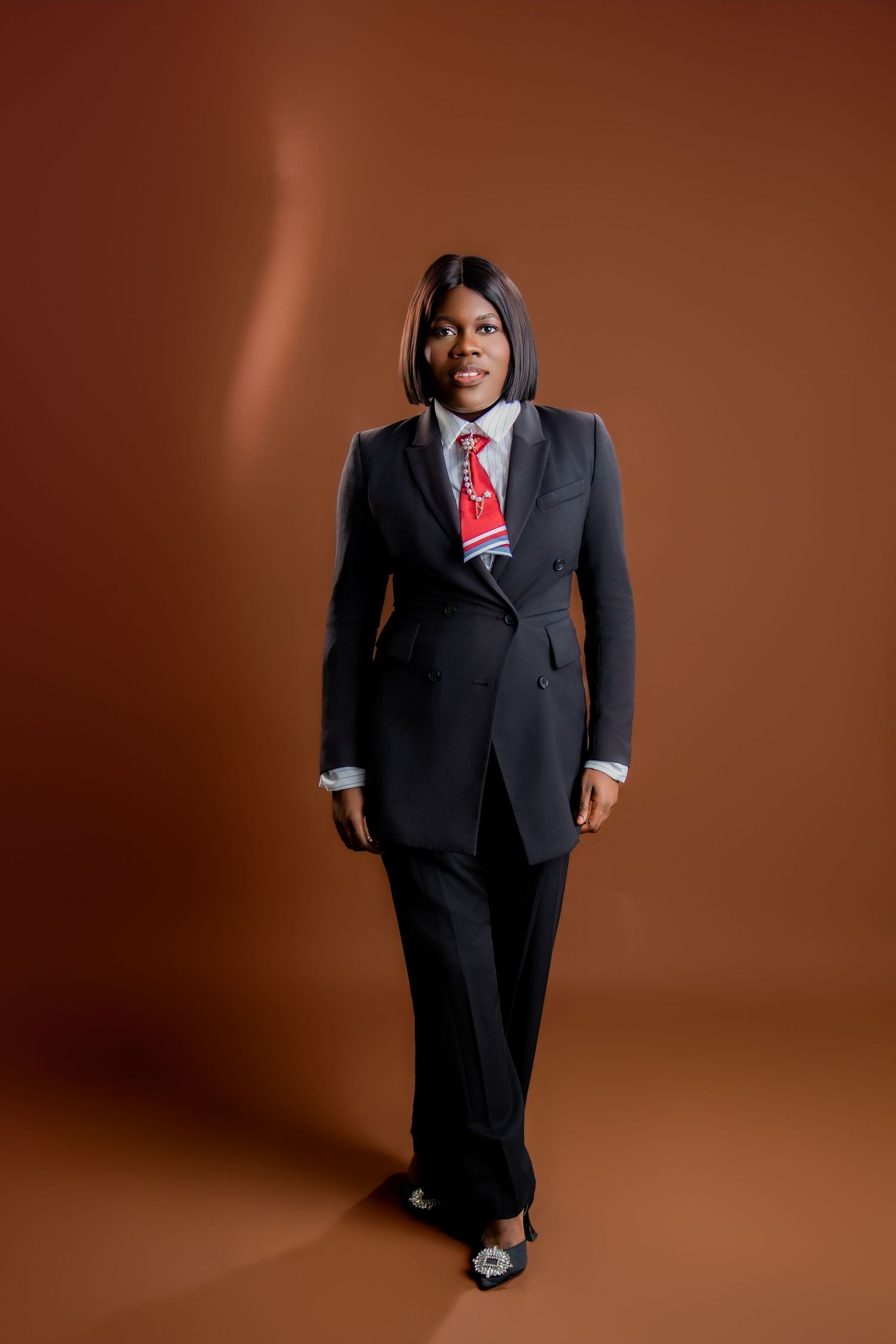 A confident Black businesswoman in a dark suit and red tie poses against a brown studio background.