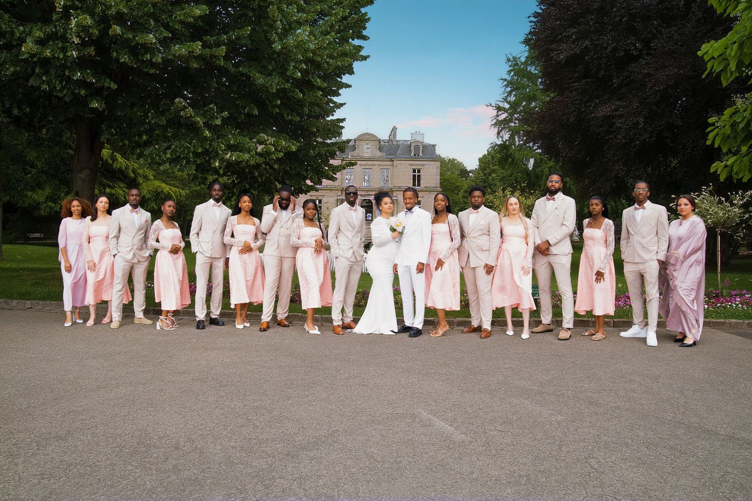 Large wedding party poses outdoors in coordinated pink and white formal attire near a grand estate.