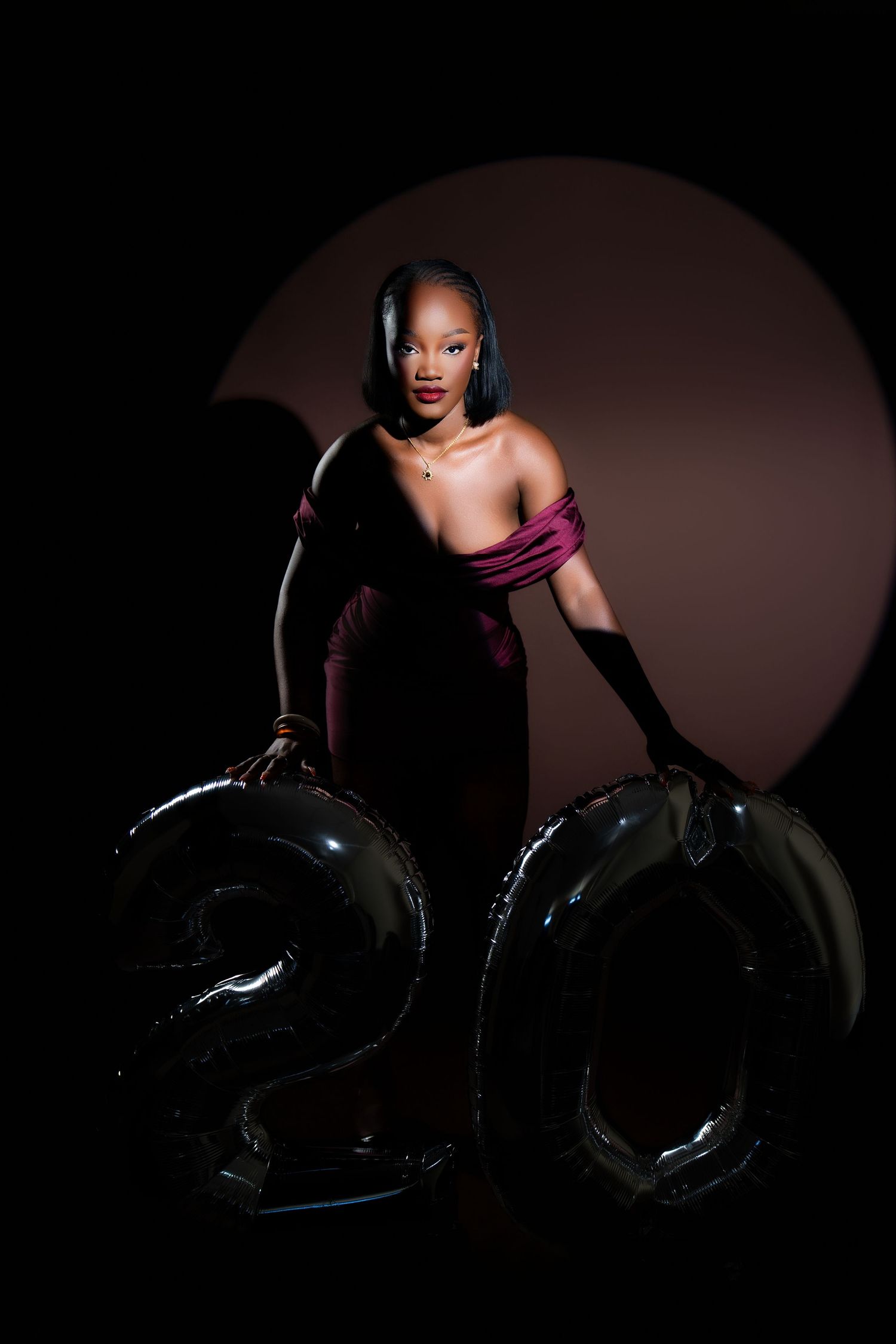 Woman in burgundy off-shoulder gown holding large black number 20 balloons against dark backdrop.