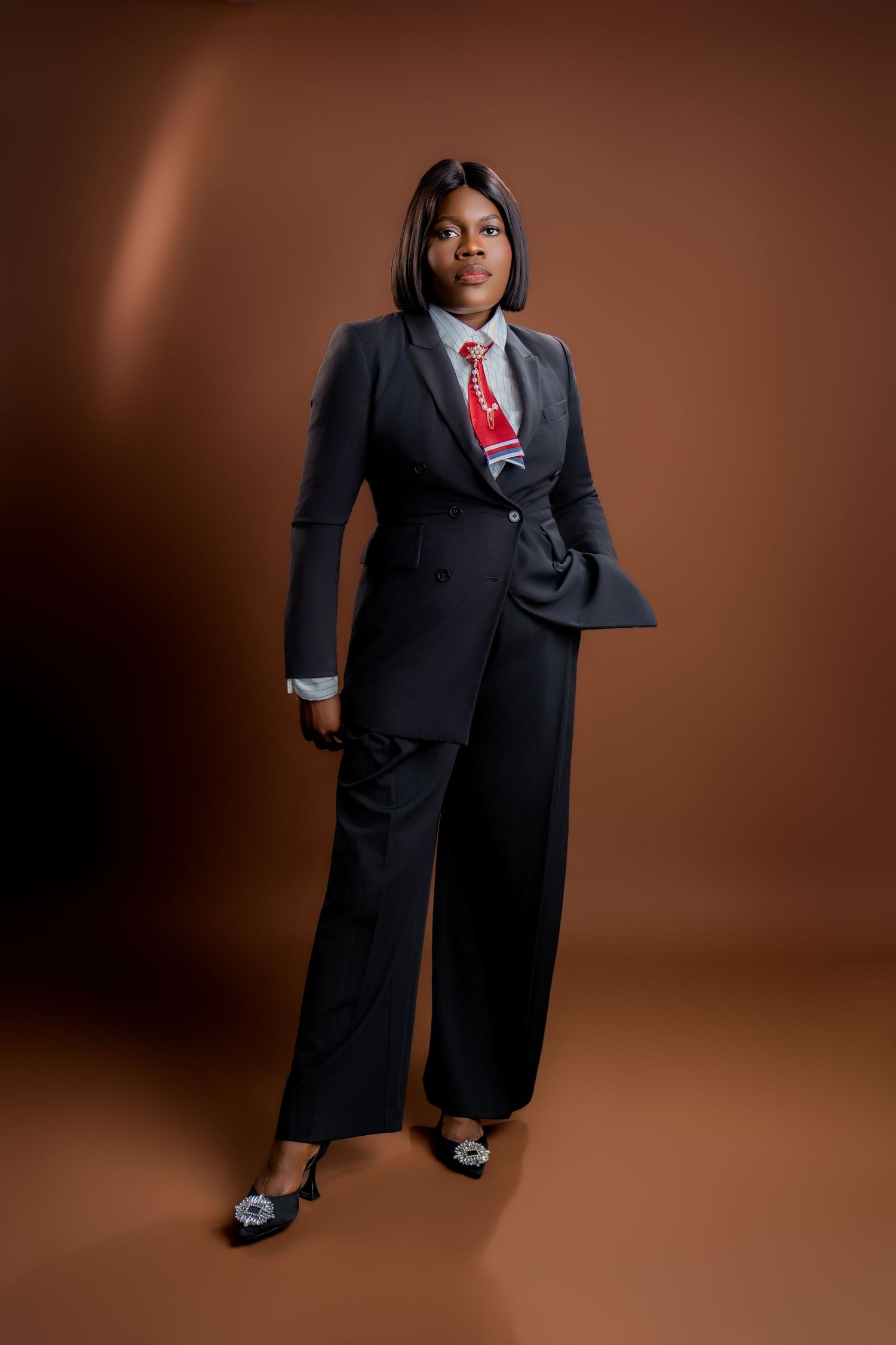 A confident Black woman in a black pantsuit with red tie poses against a brown studio background.