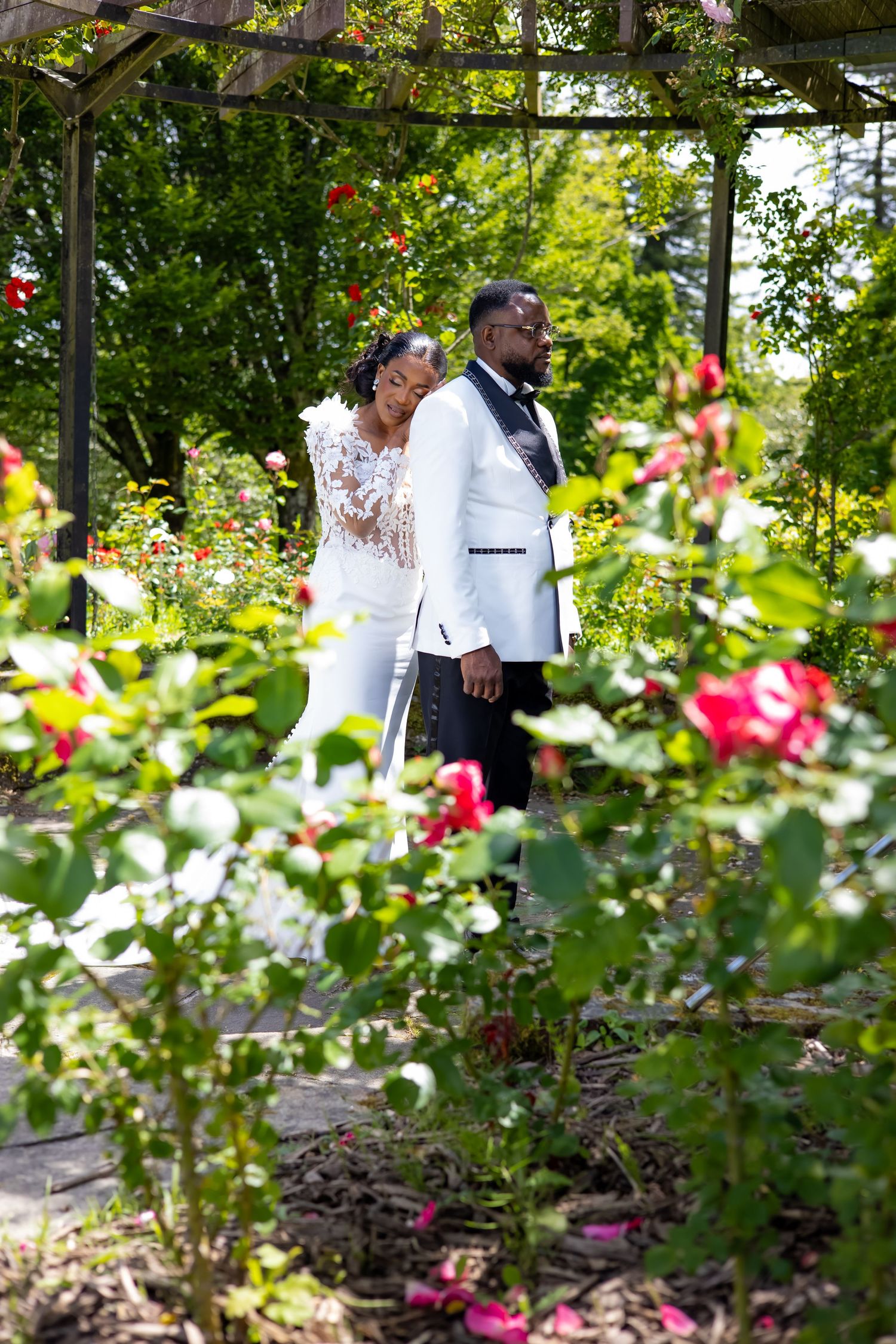 A bride and groom in white wedding attire stand among colorful roses in a lush garden arbor.