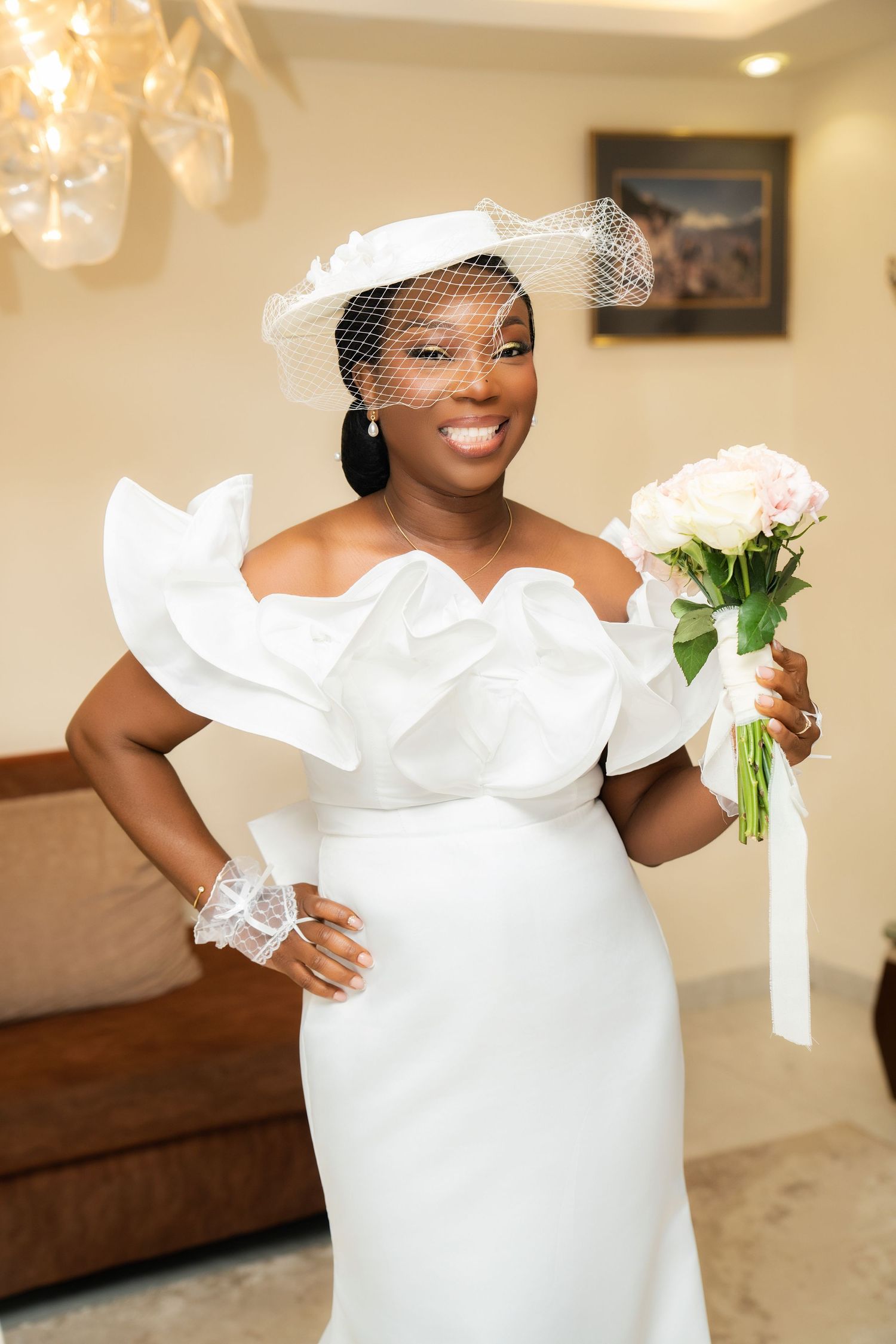 Smiling Black bride in white off-shoulder ruffled gown and birdcage hat holding pink rose bouquet indoors.