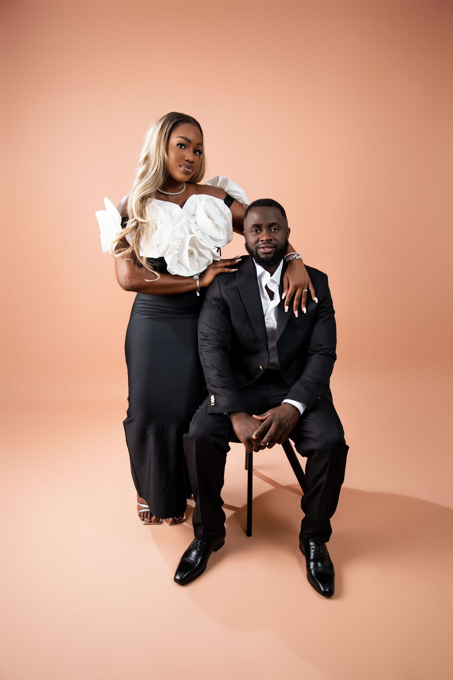 Elegant Black couple posing in studio, woman in white ruffled top and black skirt stands behind seated man in black suit.