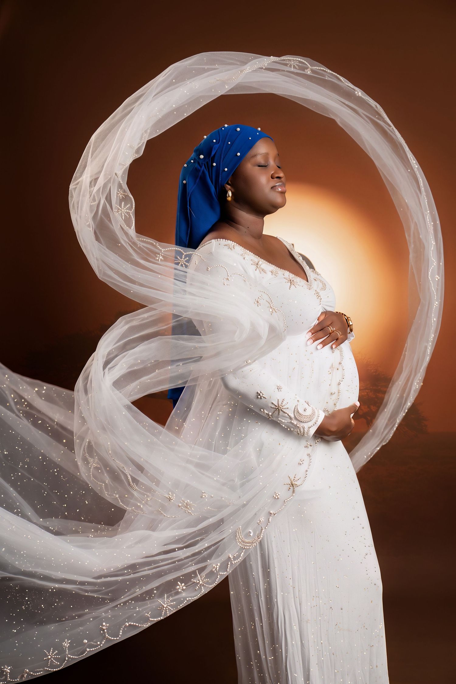 Pregnant Black woman in white embroidered gown and blue headwrap poses with flowing white veil against warm brown backdrop.