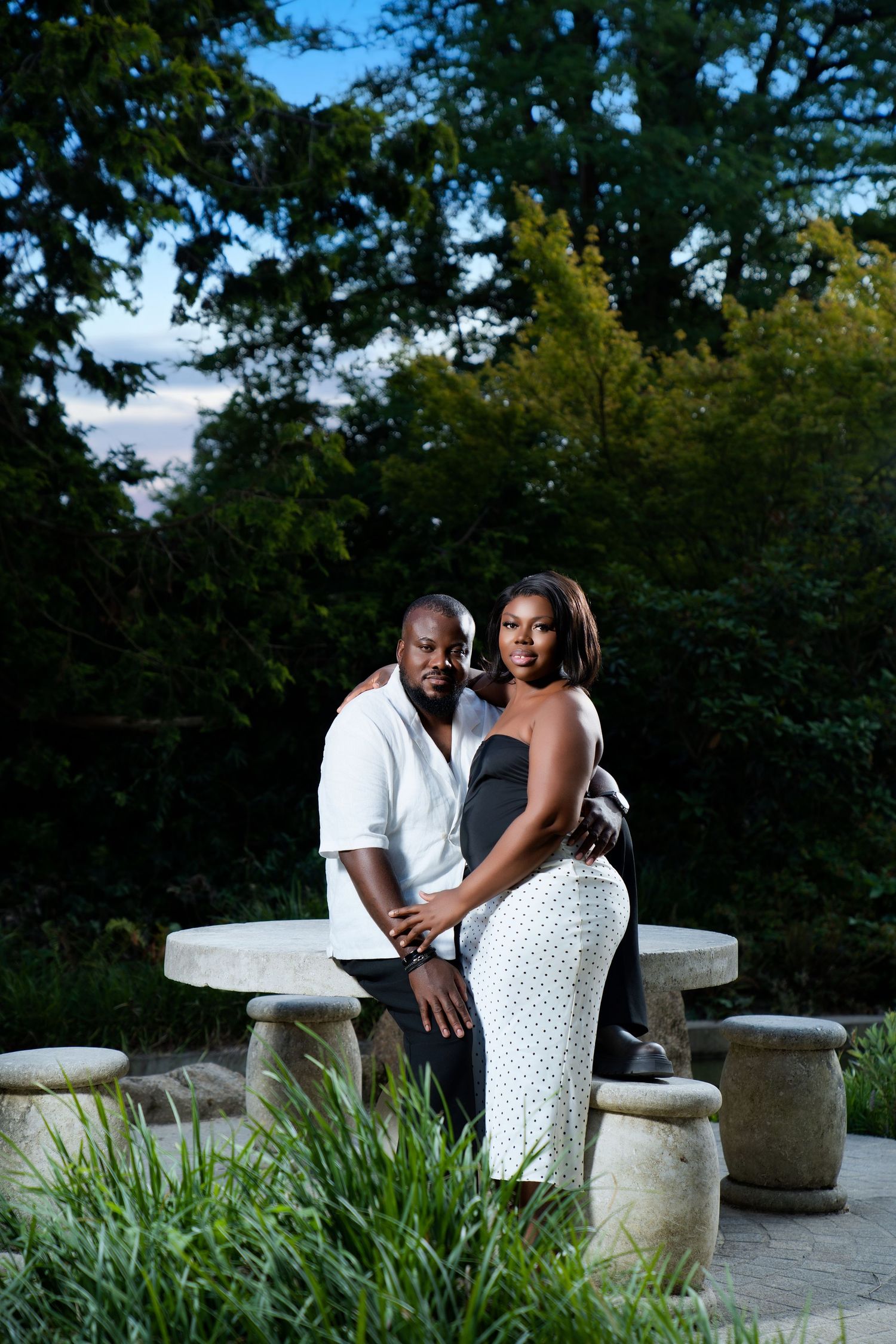 A couple poses elegantly near a stone table in a lush garden at dusk, woman in white dress, man in white shirt.