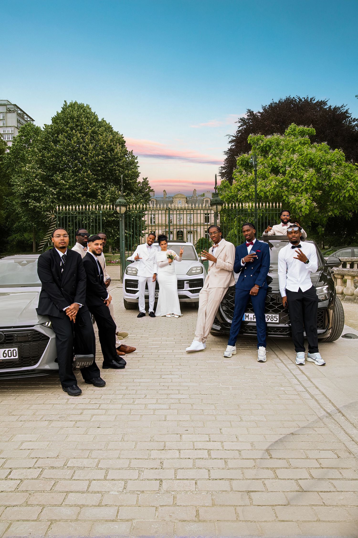 Wedding party poses outdoors near luxury cars at dusk, bride in white gown surrounded by groomsmen.