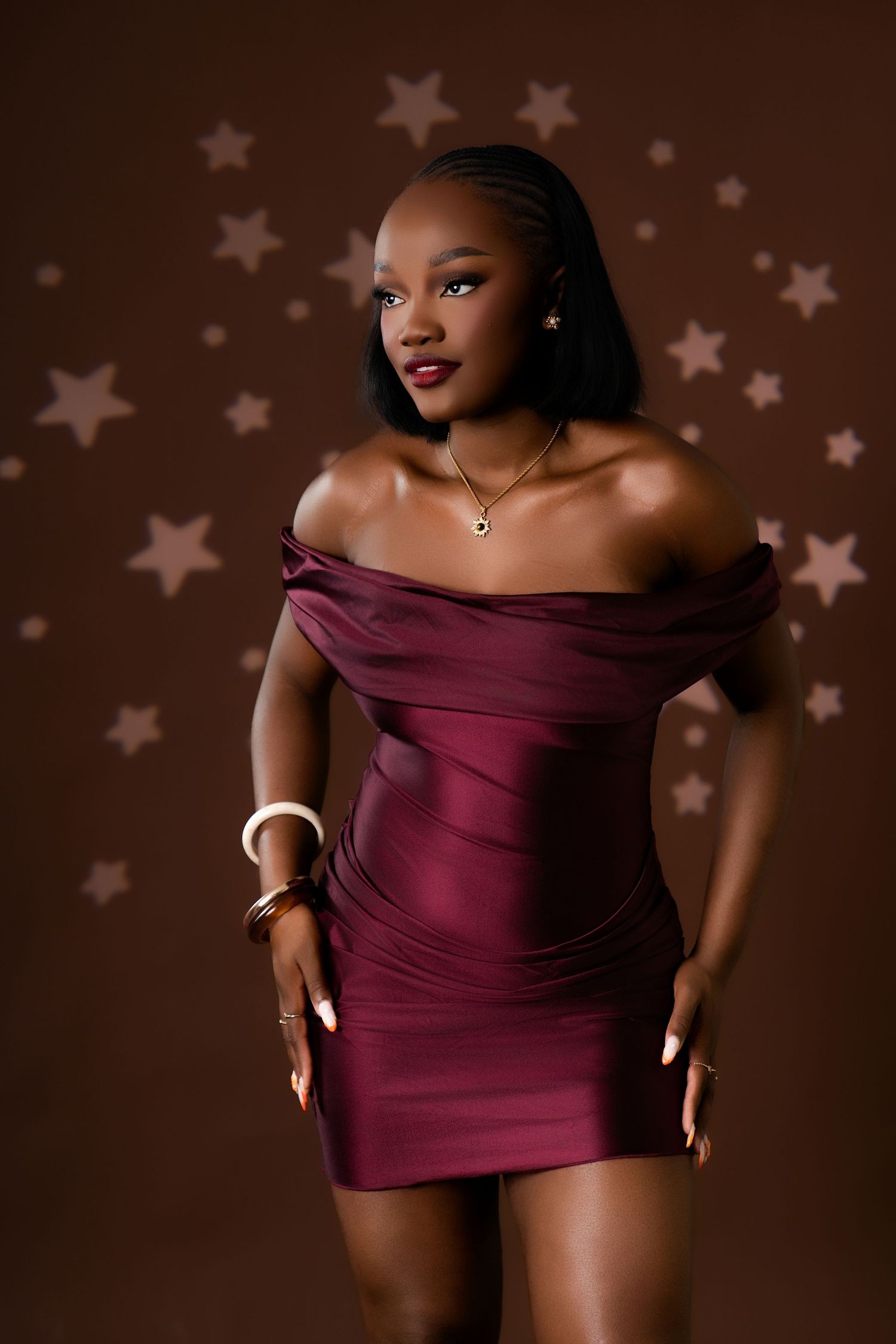 Young woman in a burgundy off-shoulder mini dress poses against a brown backdrop with star decorations.