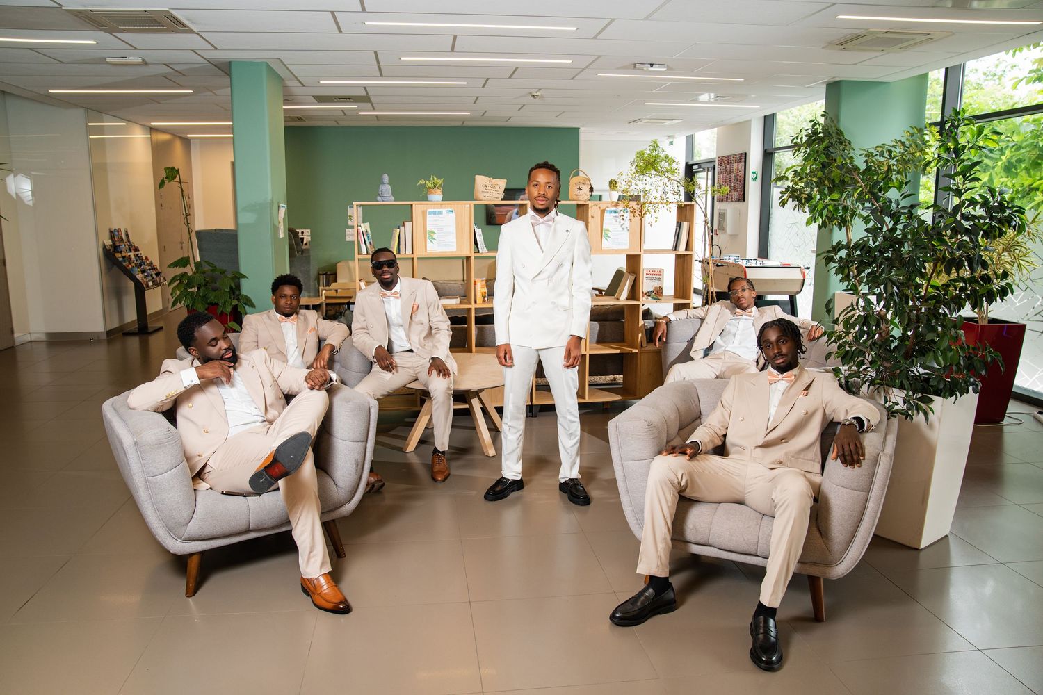 Six well-dressed Black men in cream suits pose elegantly in a modern office lobby for a formal group photo.