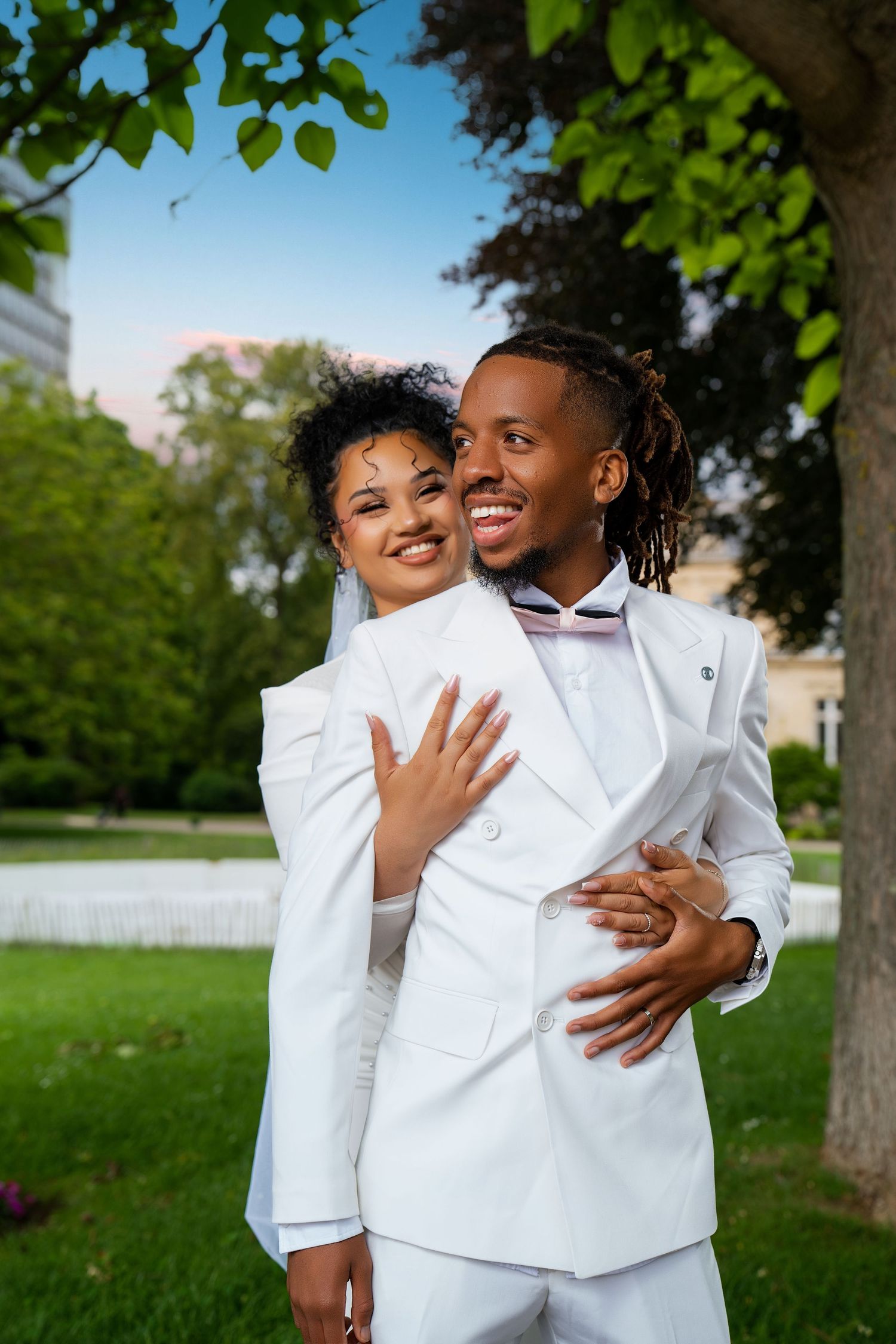 A joyful Black couple in matching white wedding attire embrace outdoors, smiling in a lush green park setting.