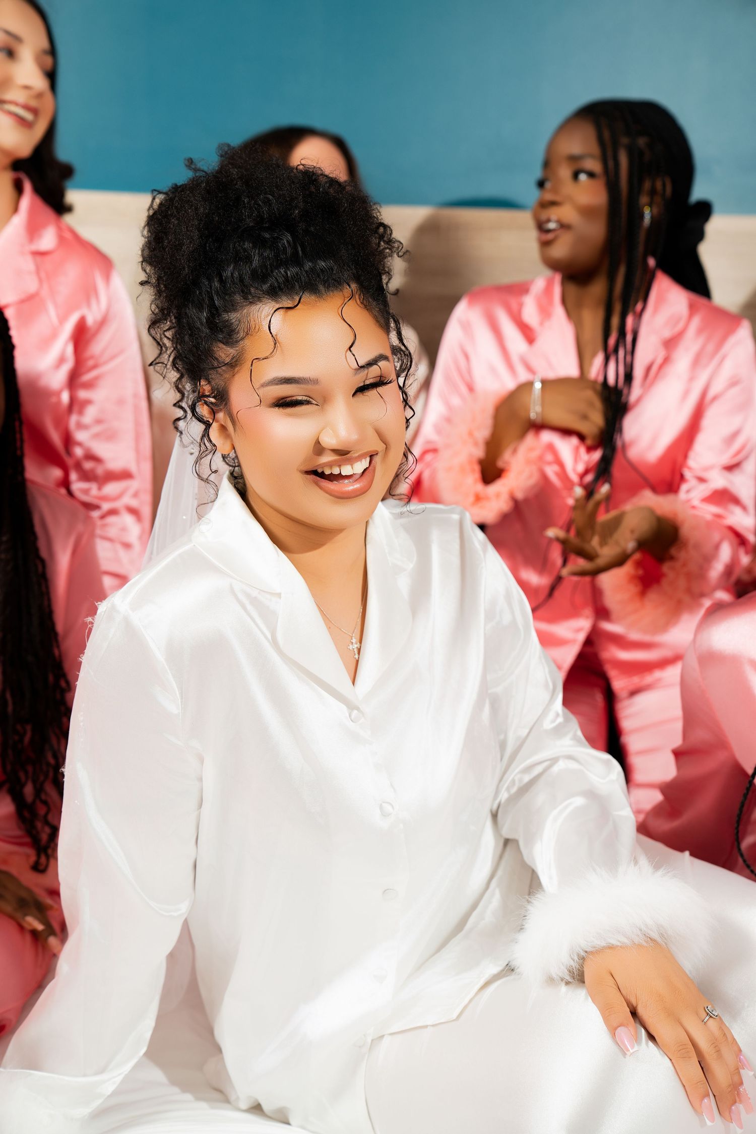 Smiling bride in white satin pajamas with bridesmaids in pink satin on a bed during bridal party preparations.