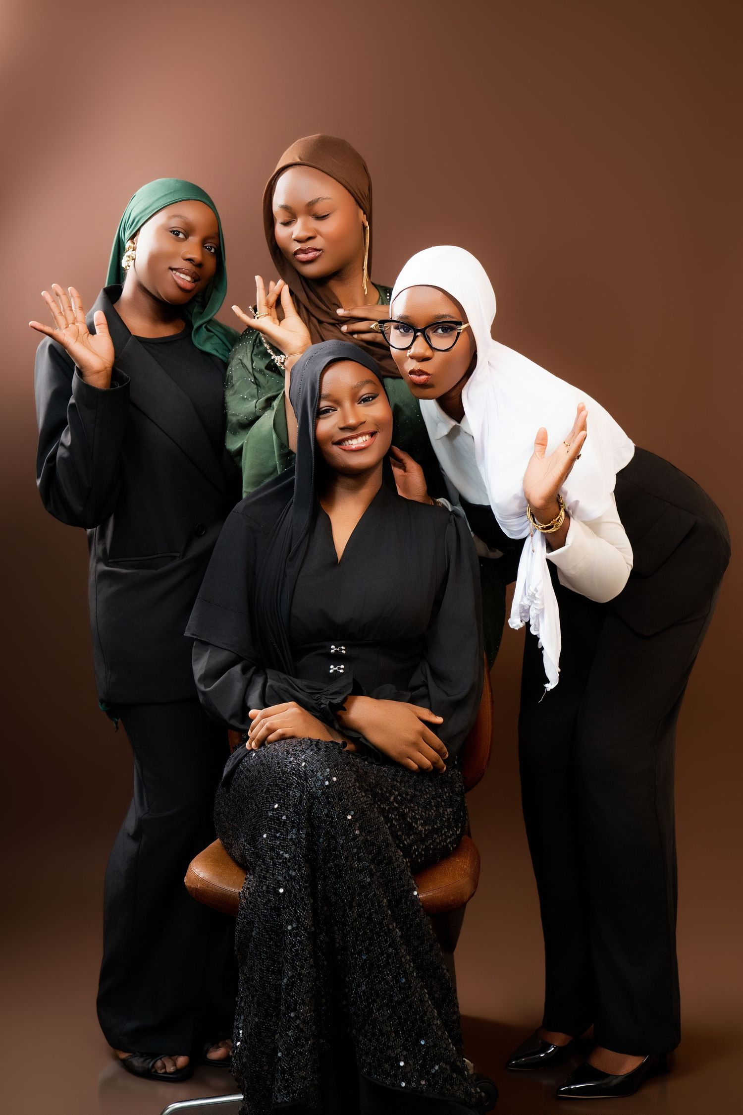 Four Muslim women in hijabs pose together in a studio, wearing elegant black outfits against a brown background.
