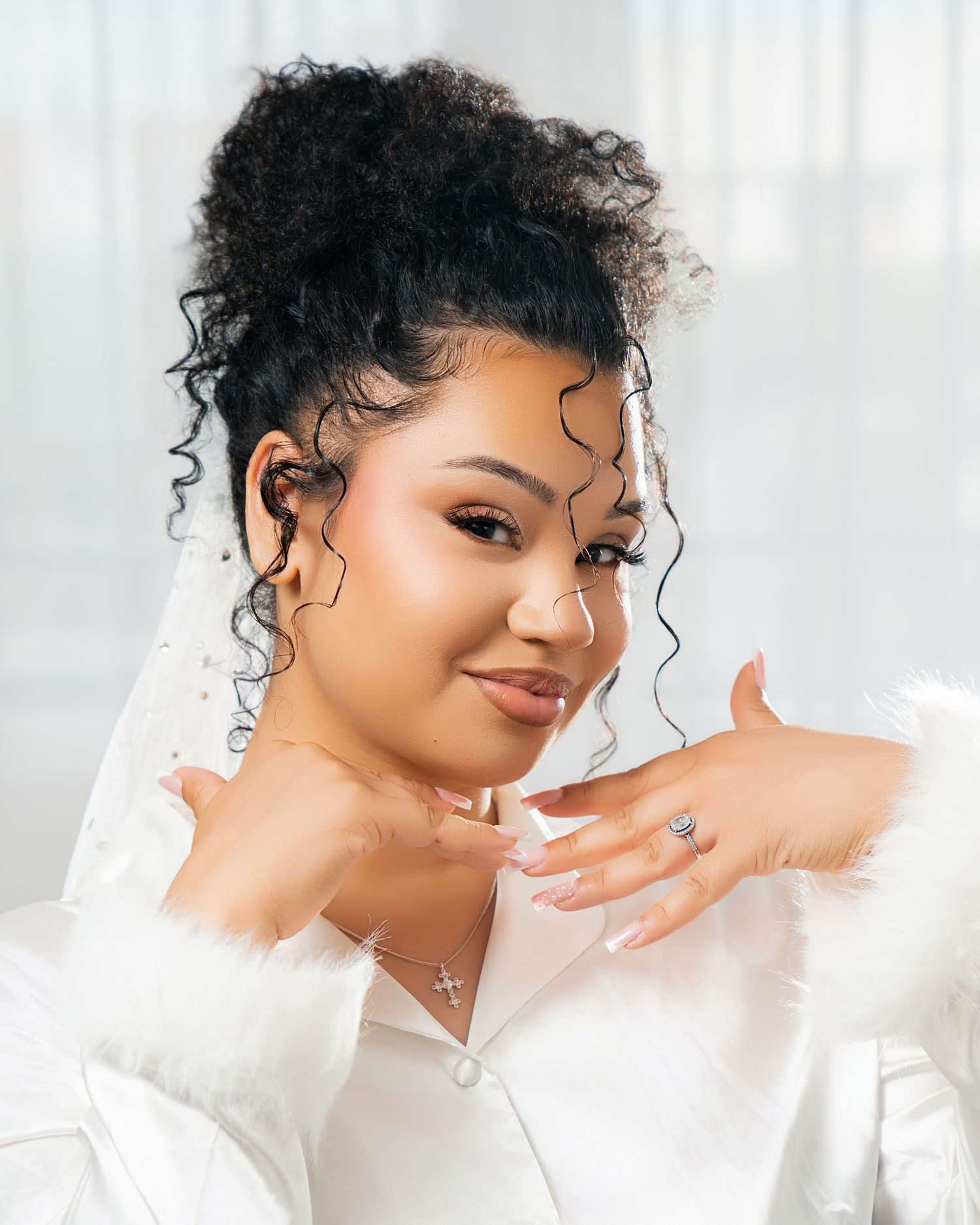 Bride in white robe with curly updo, veil, and ring posing elegantly.