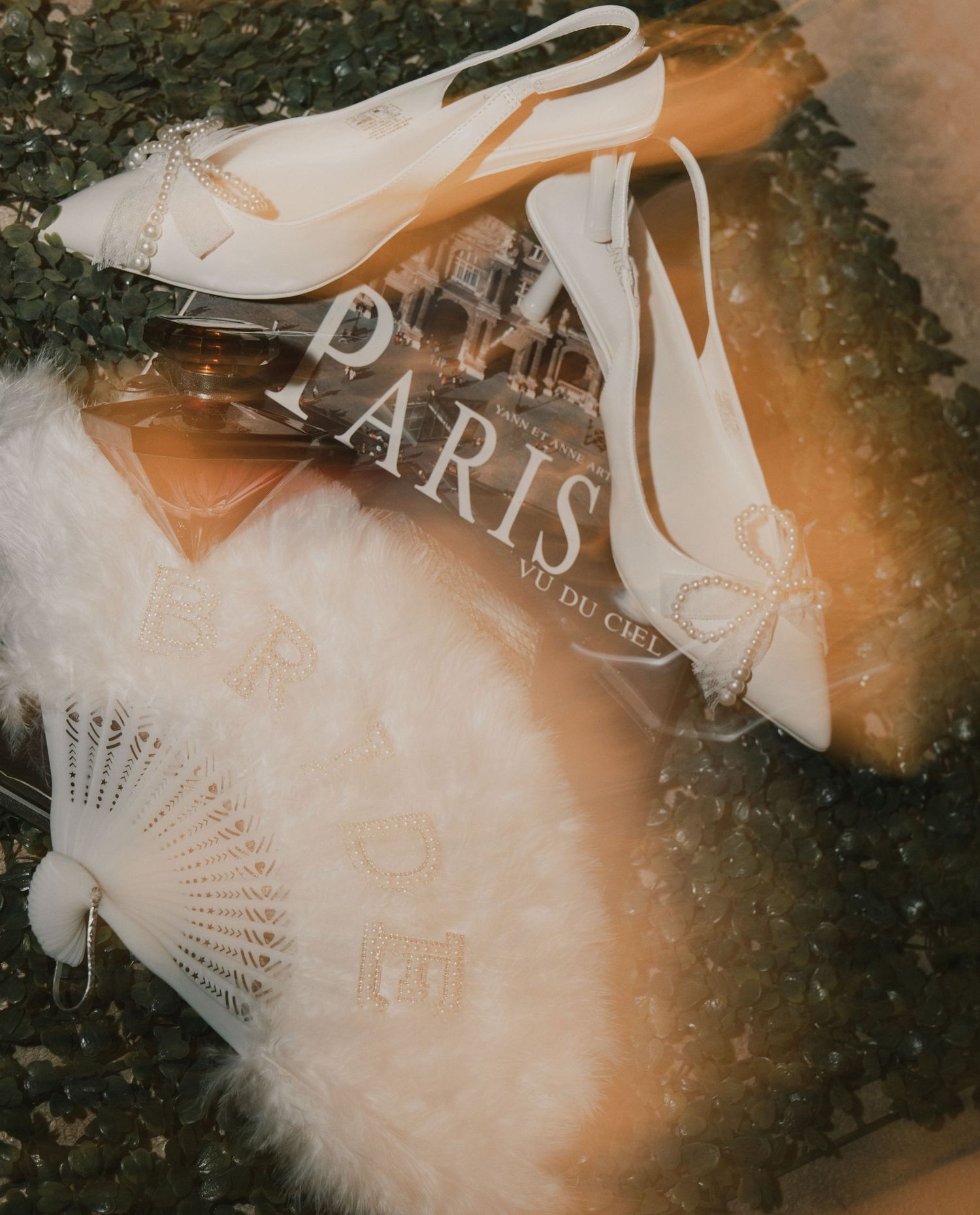 Elegant bridal flat lay featuring white heels, a decorative fan, and a Paris book on a textured surface.