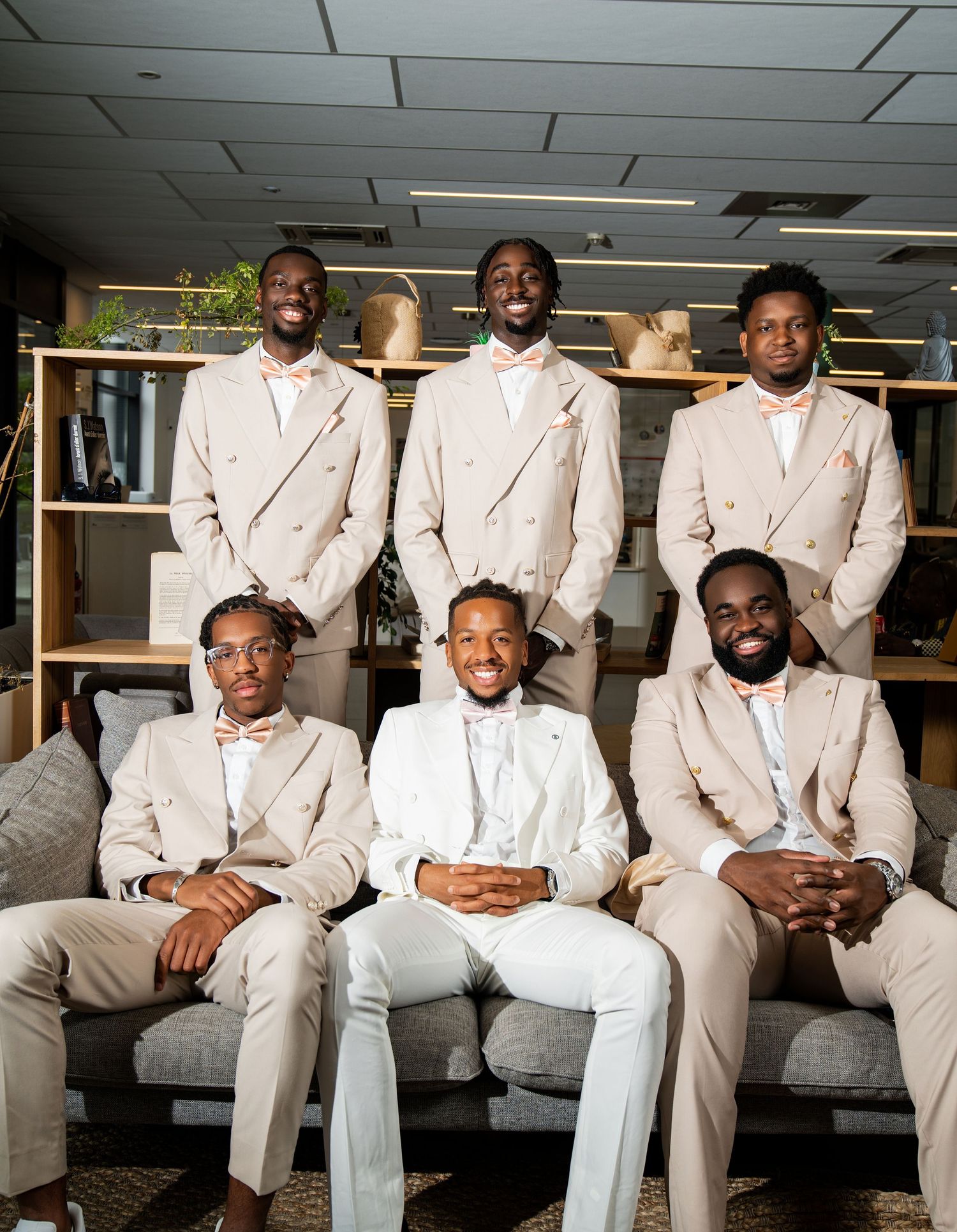 Six men in matching beige suits and bow ties pose together, with one man in white seated at center.