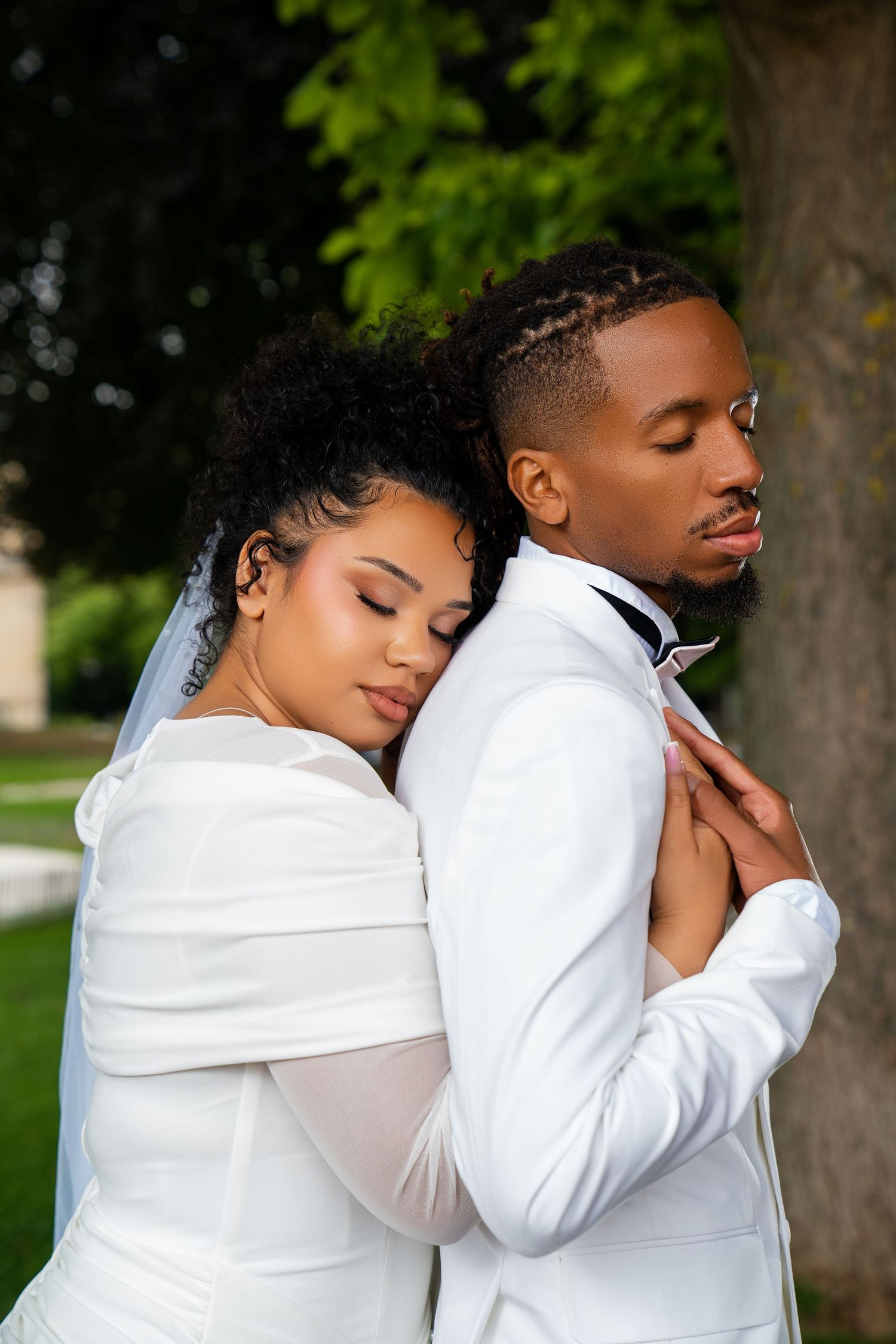 Black bride in white gown embraces groom in white suit outdoors near a tree in a romantic wedding portrait.