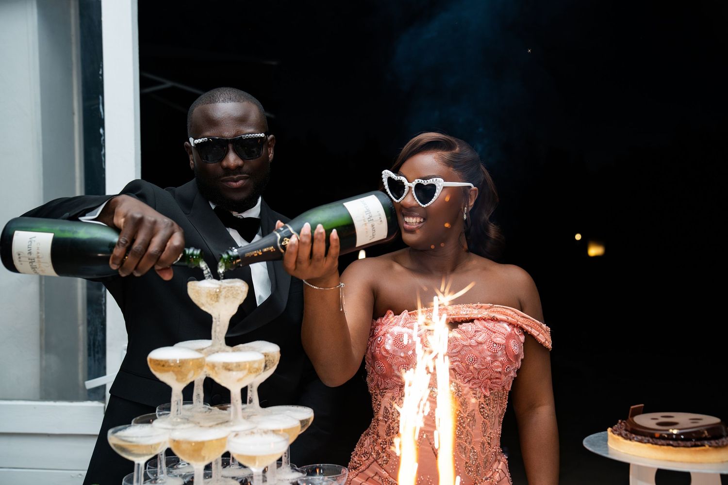 Couple pouring champagne tower at celebration, woman in pink dress with heart sunglasses, man in suit holding bottle.
