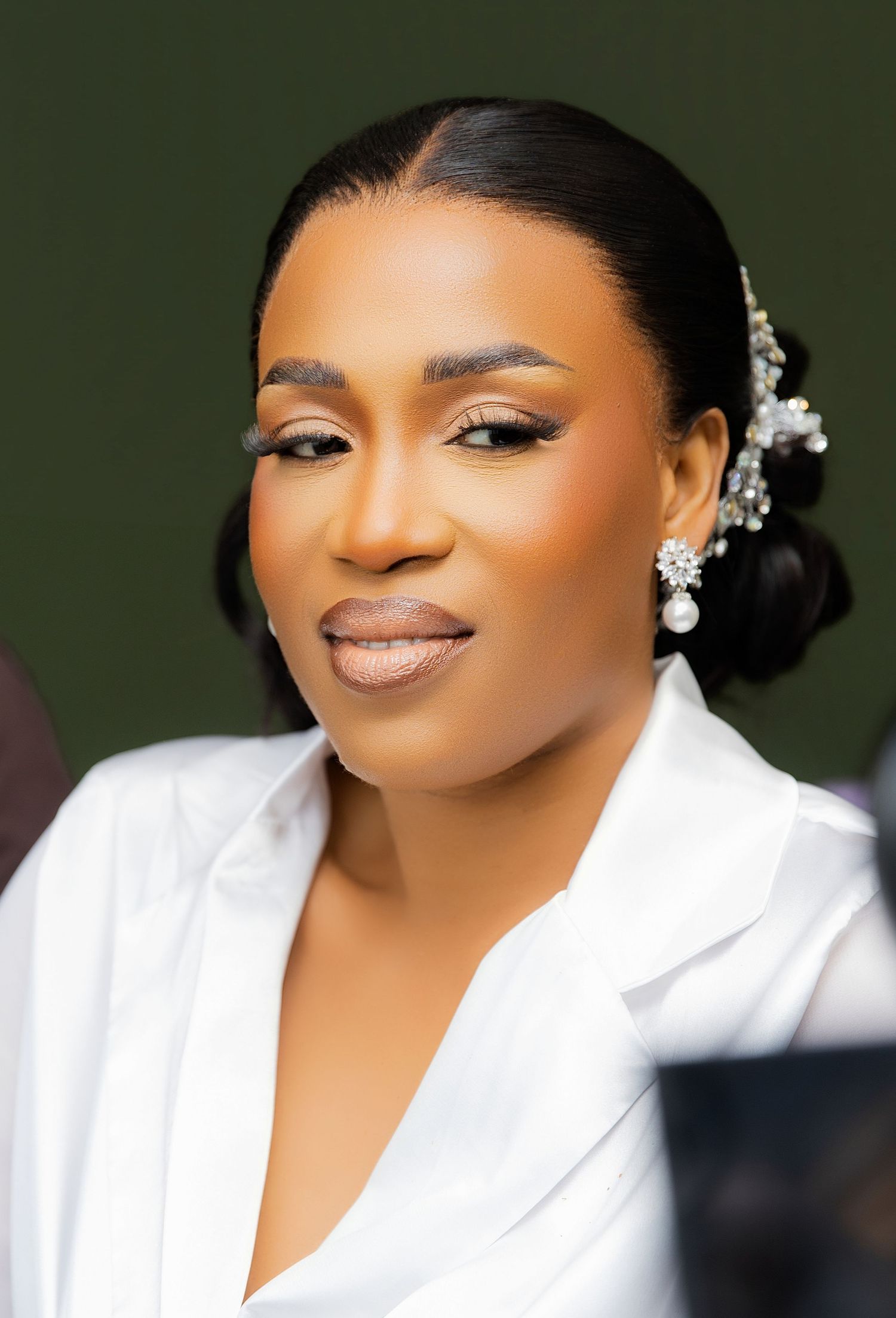 Elegant woman with bridal updo, crystal hair accessories, diamond earrings, and flawless makeup in a white outfit.