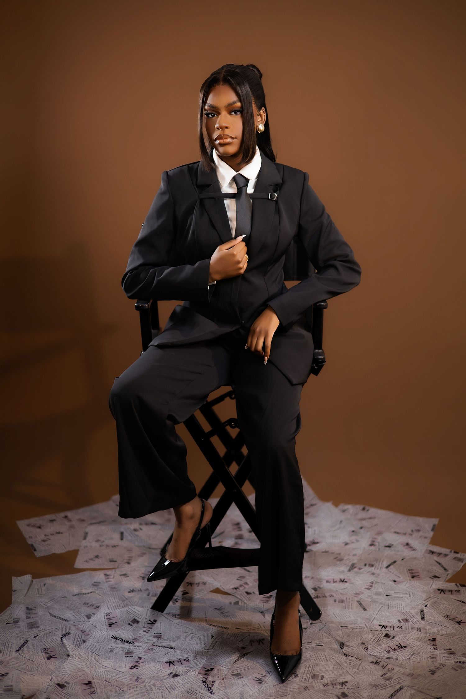 A confident person in a black suit sits in a director's chair surrounded by papers on a brown studio background.