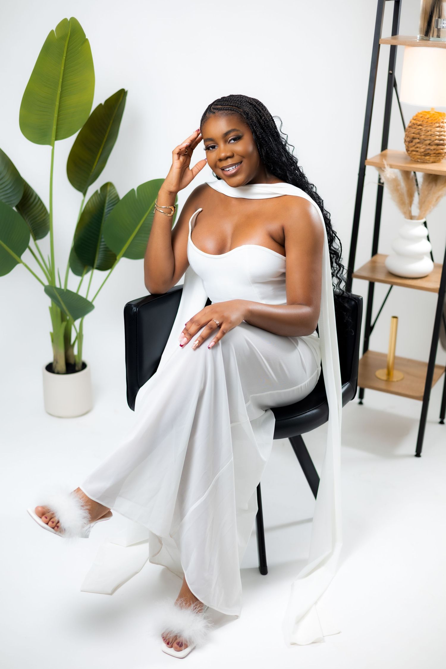 A woman in a white jumpsuit and fluffy slides sits gracefully in a black chair, posing in a bright studio setting.