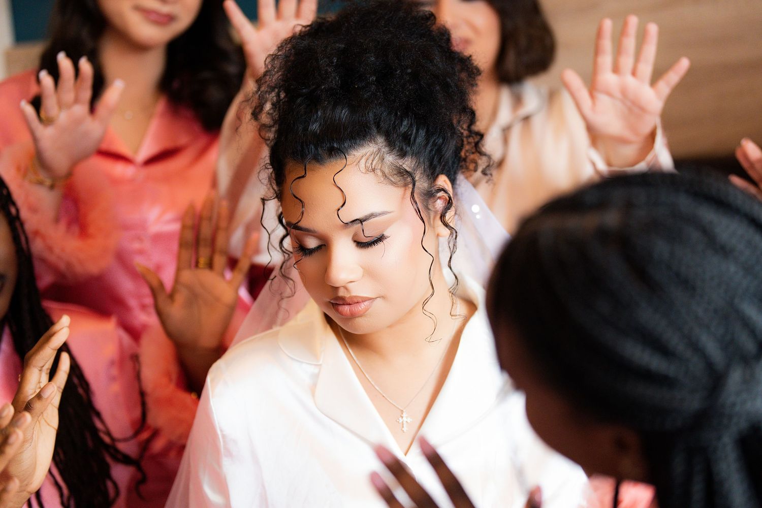 Bride in white robe surrounded by bridesmaids raising hands in celebration during wedding morning preparations.