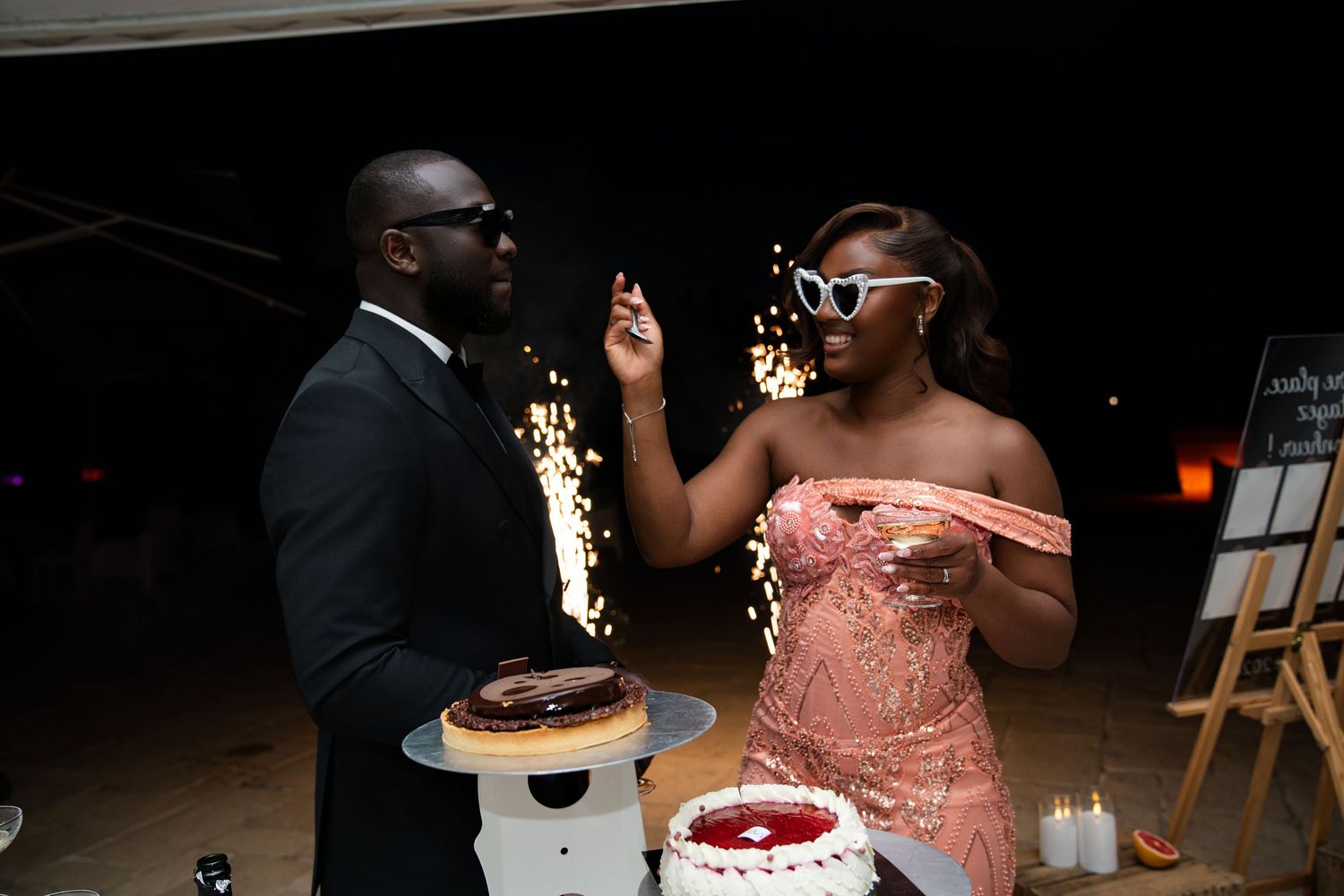 Couple celebrating at night event, man in black suit and woman in pink dress cutting cake with sparklers.