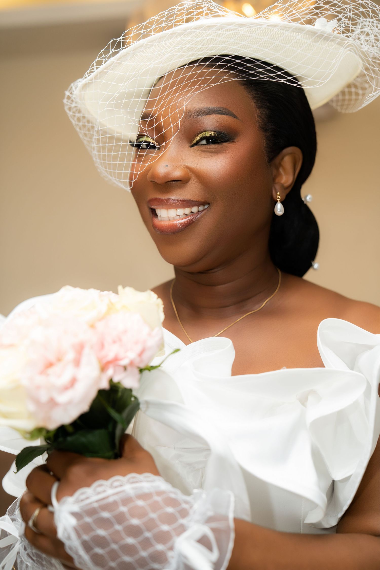 Smiling Black bride in white off-shoulder gown and veiled hat holds pink roses bouquet on her wedding day.