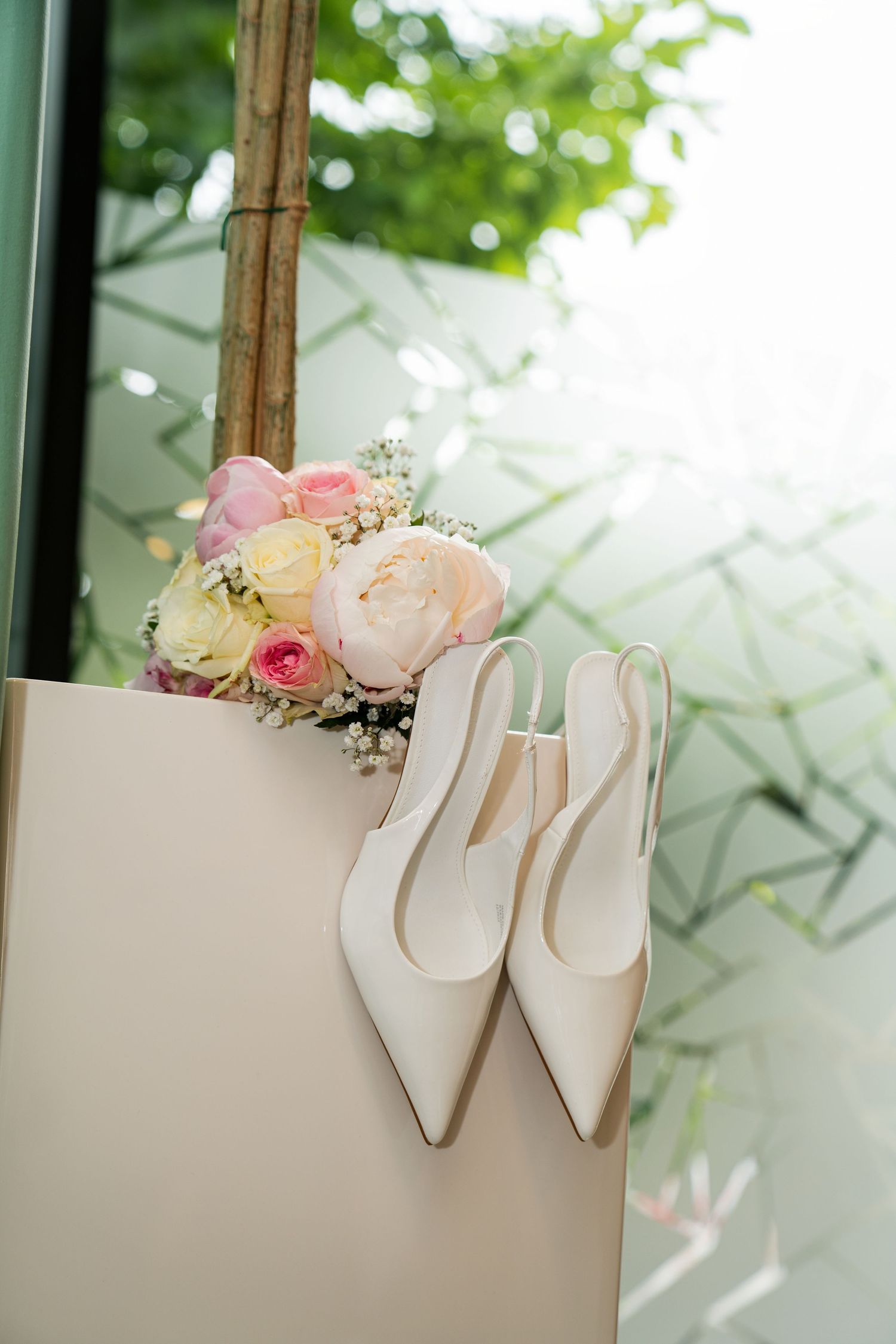 White bridal heels and pastel flower bouquet with pink and cream roses displayed on a white surface near a window.