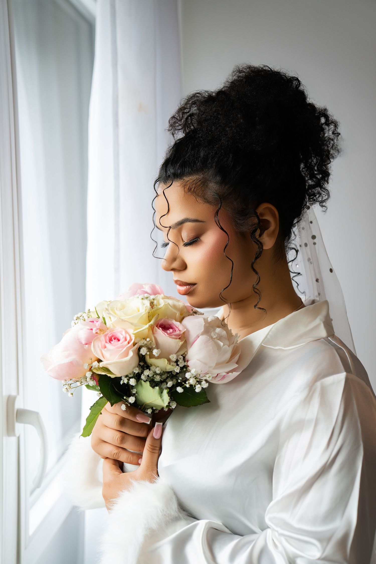 A beautiful African American bride in white dress holds pink roses bouquet near window on her wedding day.