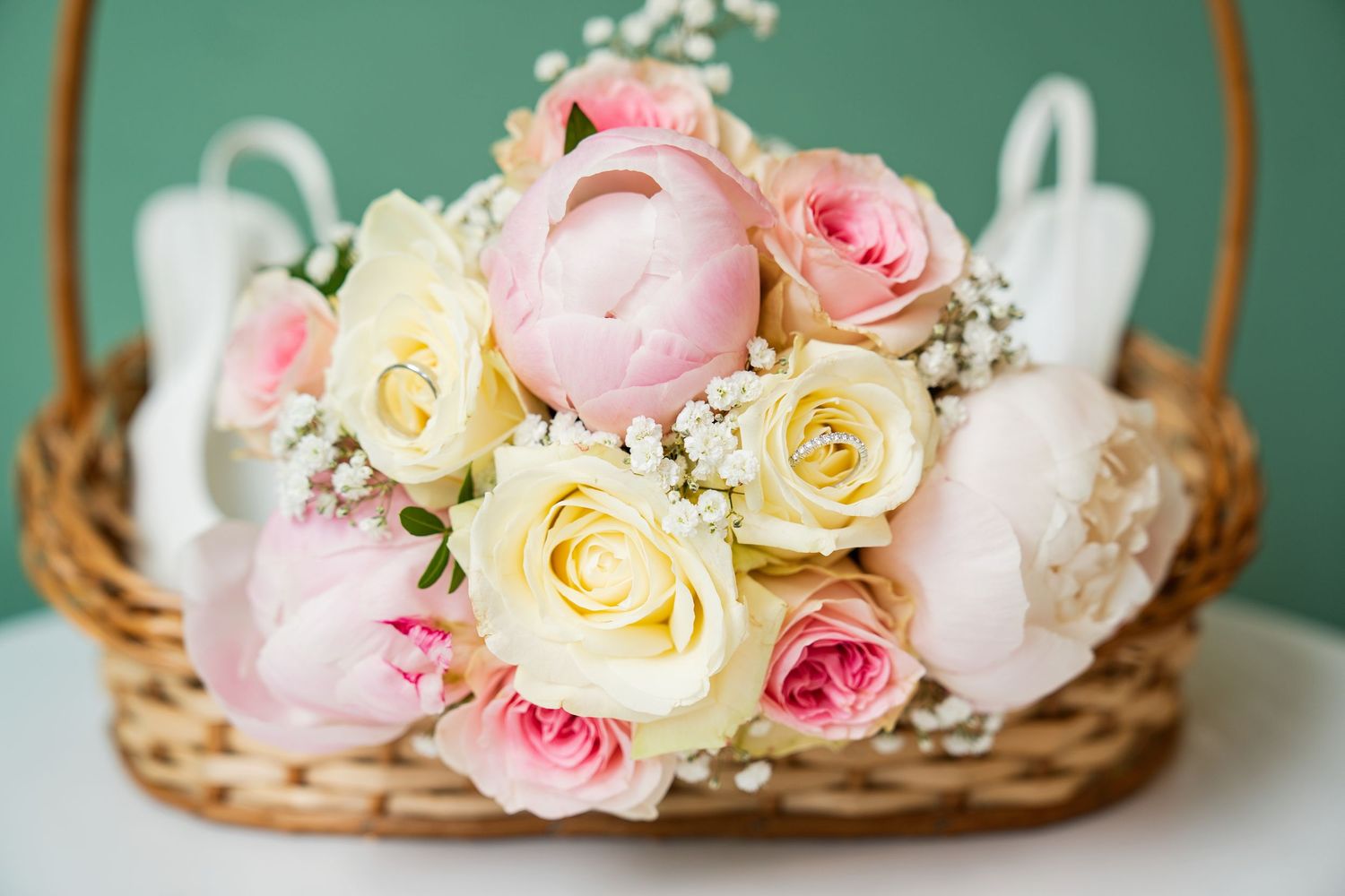 Wicker basket filled with pink peonies, cream roses, and baby's breath on a white surface with green background.