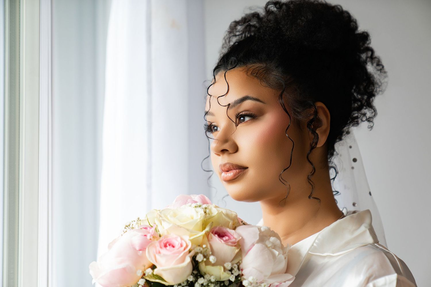 Bride in white dress holding pink and white rose bouquet, gazing out window with veil and elegant updo hairstyle.
