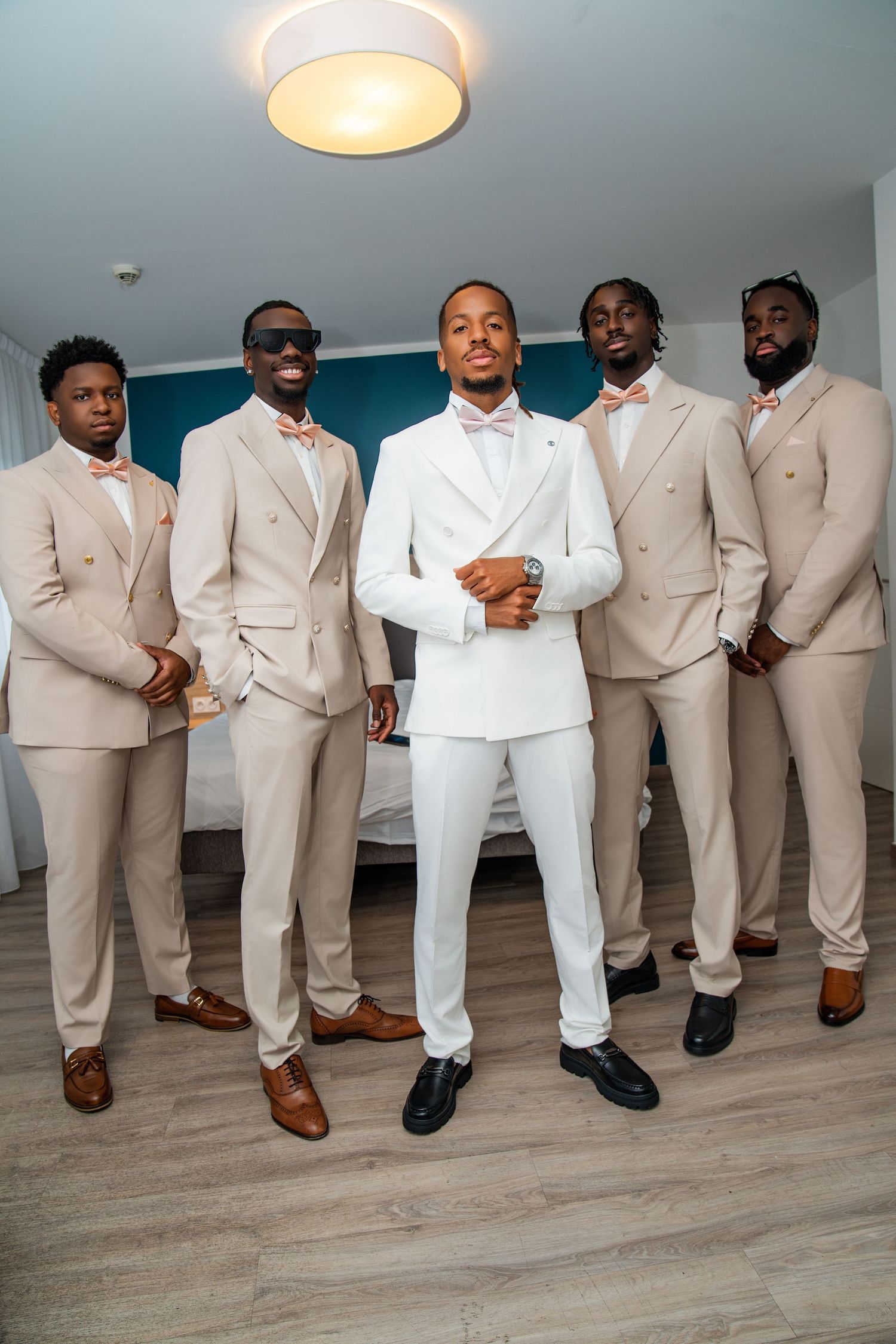 Groom in white suit poses with four groomsmen in beige suits and pink bow ties in a modern bedroom.