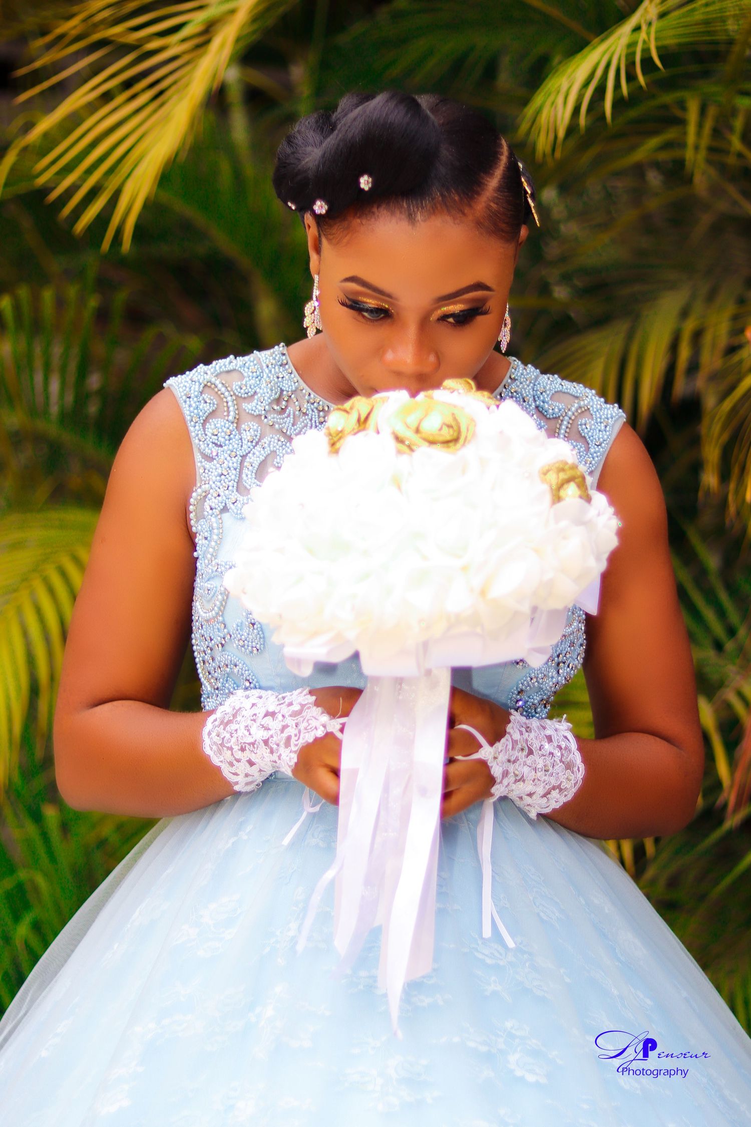 Bride in delicate lace gown poses with white bouquet against tropical palm background.
